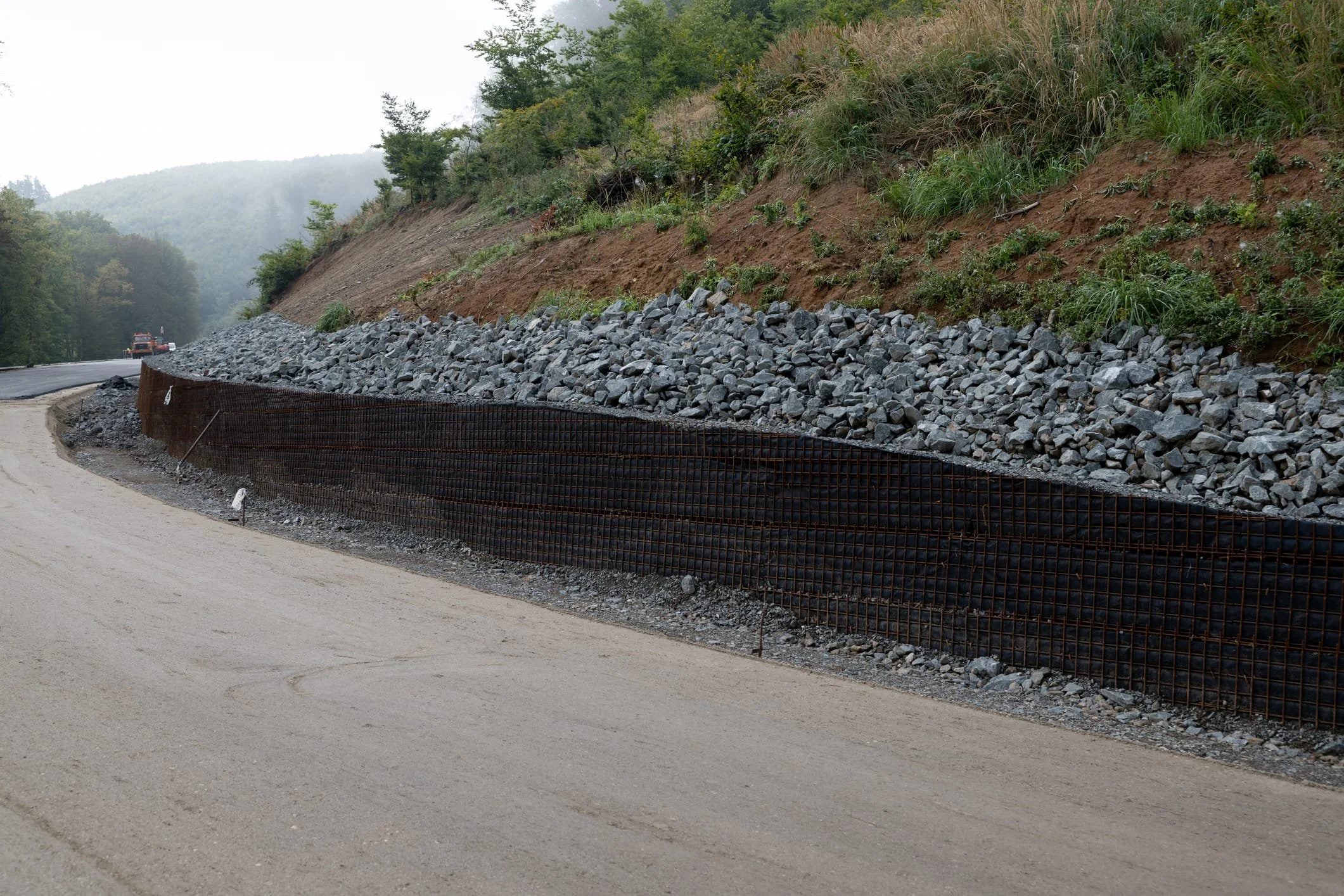A road under construction with gravel and a temporary metal barrier on the side, set against a hillside with trees and vegetation.