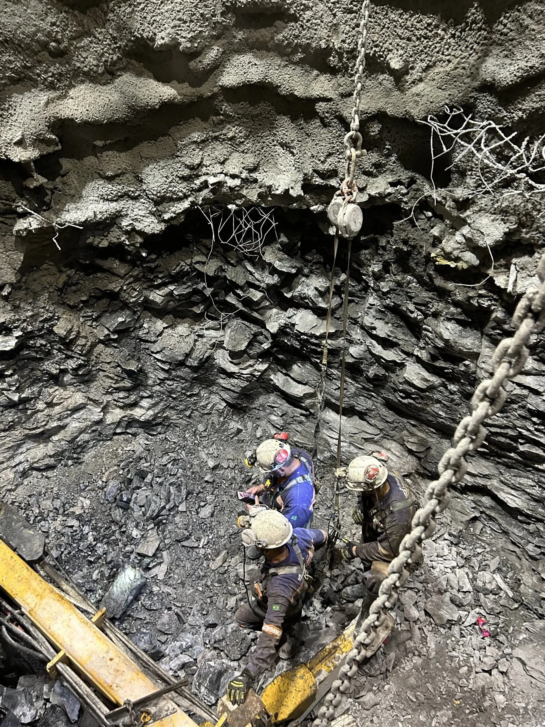 Mining workers wearing helmets and safety gear in a dark, rocky underground tunnel, surrounded by loose rocks and a large chain hanging from the ceiling.