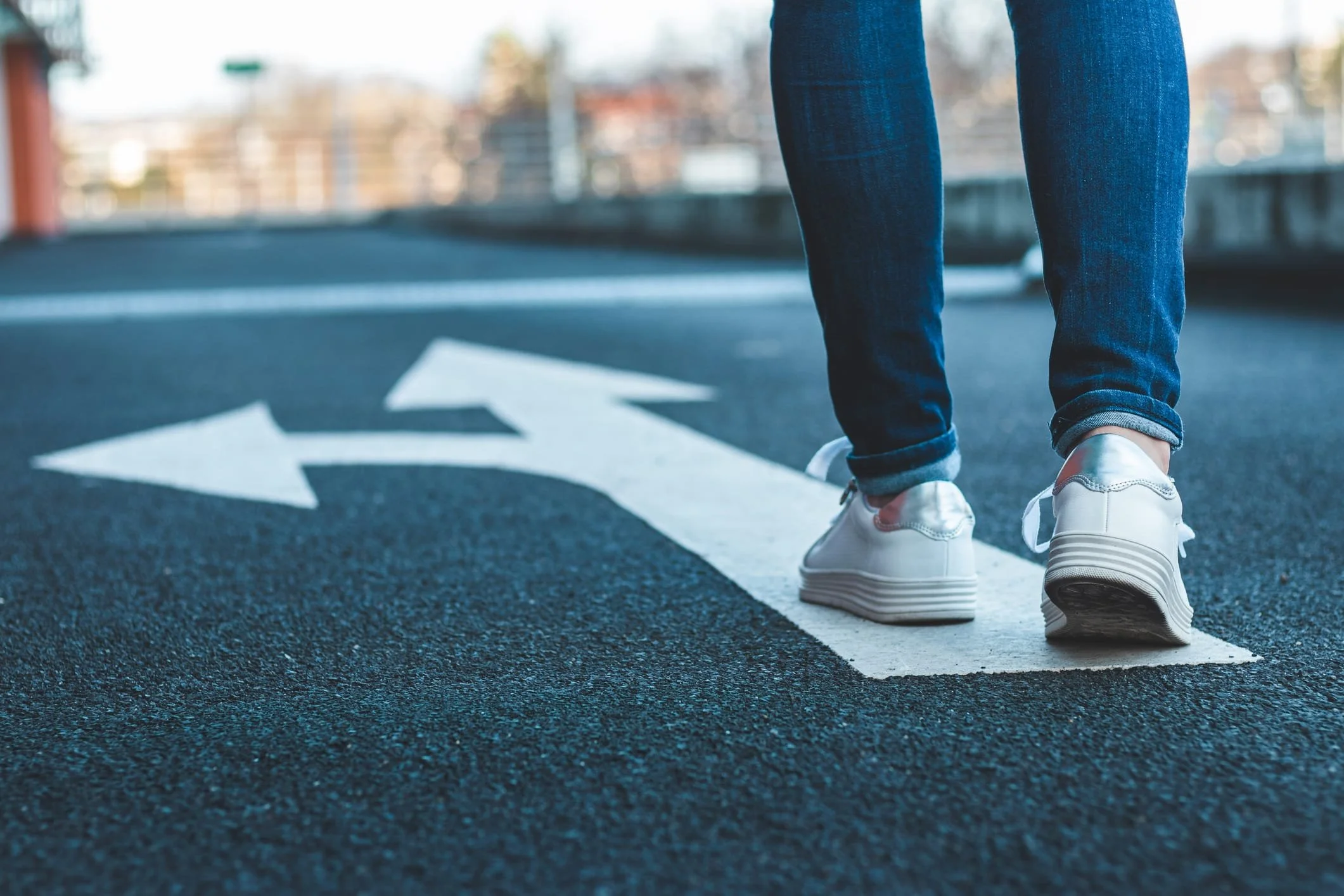 A person wearing white sneakers and blue jeans standing on a street with a white arrow painted on the road.