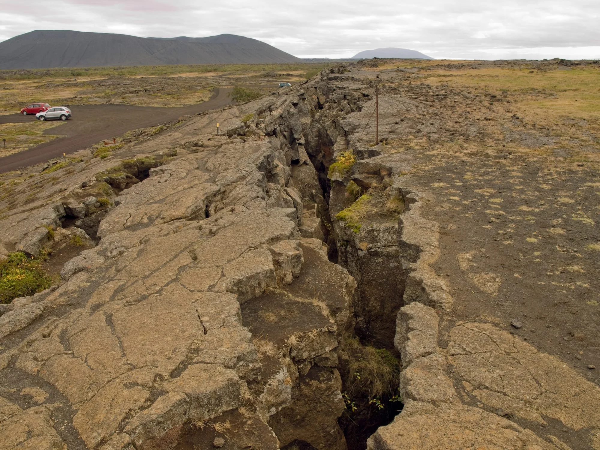 A cracked and broken volcanic rock formation in a barren landscape with a dirt road and cars parked nearby. In the background, there are hills or mountains under a cloudy sky.