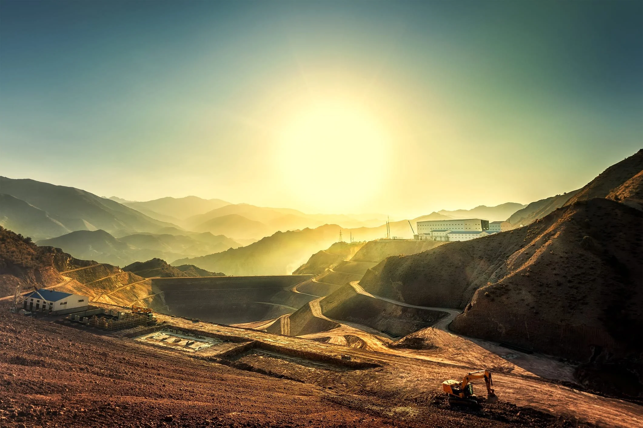 Sunset over a mountainous landscape with winding roads, construction equipment, and a building under construction.