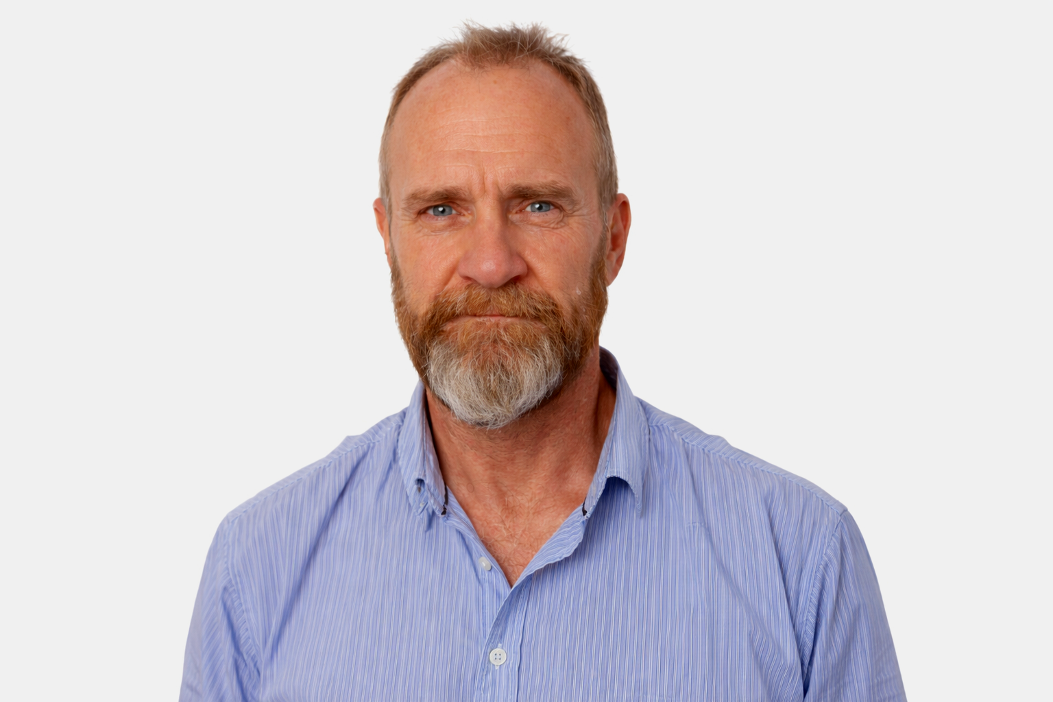Close-up portrait of a middle-aged man with light skin, gray hair, and a beard, wearing a blue striped shirt, against a black background.