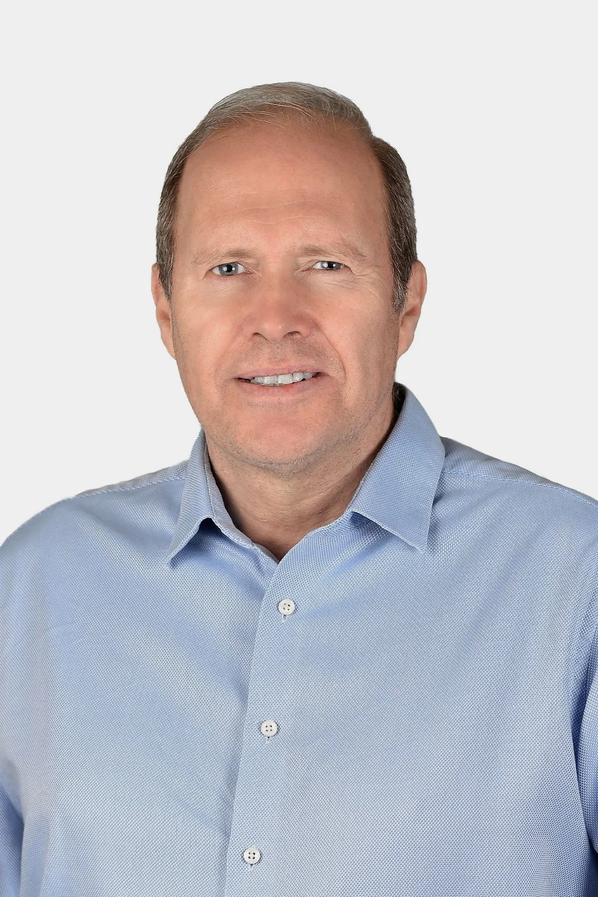 A man with short hair wearing a dark blazer and light shirt, standing outdoors with green foliage in the background.