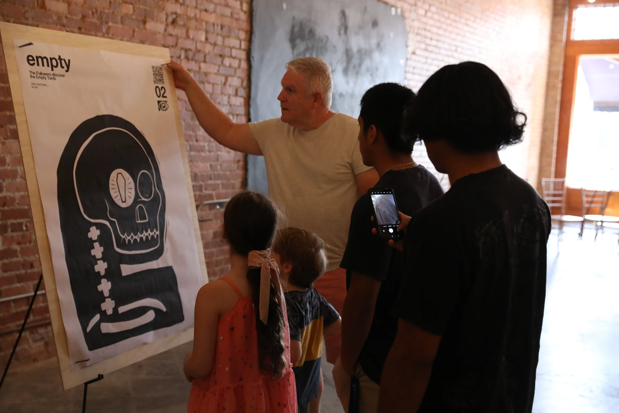 A man is giving a presentation on an art exhibit to a group of children and teenagers, with the focus on a large poster of a skull with a molecular structure and text that reads 'empty' and 'The Followers discover the Empty Turtle.'
