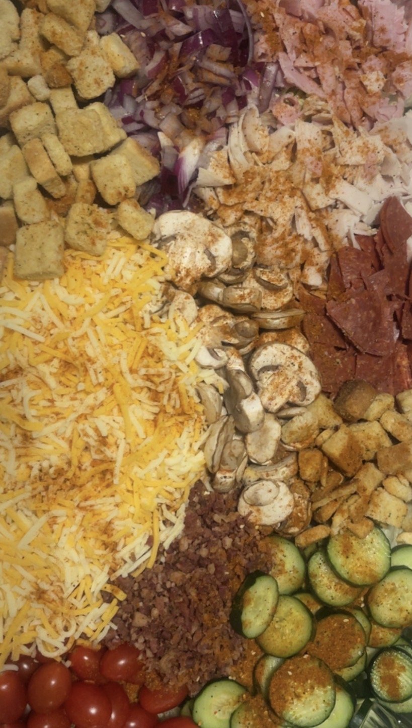 A variety of chopped ingredients for a taco bar, including cherry tomatoes, shredded cheese, sliced zucchini, cooked ground beef, diced seasoned chicken, sliced mushrooms, sliced turkey, croutons, chopped onions, and sliced sausages.
