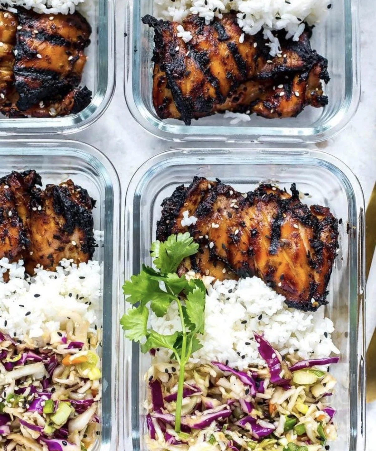 Four glass meal prep containers with grilled chicken, cooked white rice, and vegetable salad with purple cabbage and green onions, garnished with cilantro, on a light-colored surface.