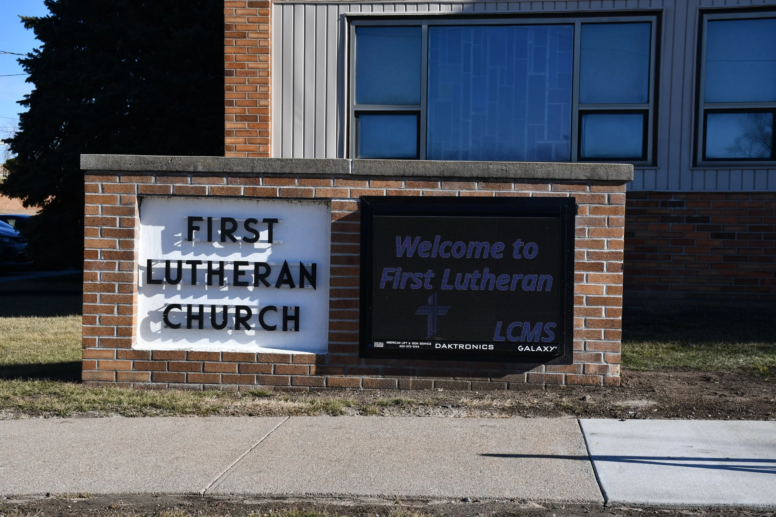 Sign on a brick wall displaying the name 'First Lutheran Church' with an electronic message board reading 'Welcome to First Lutheran LCMS' with a cross symbol.