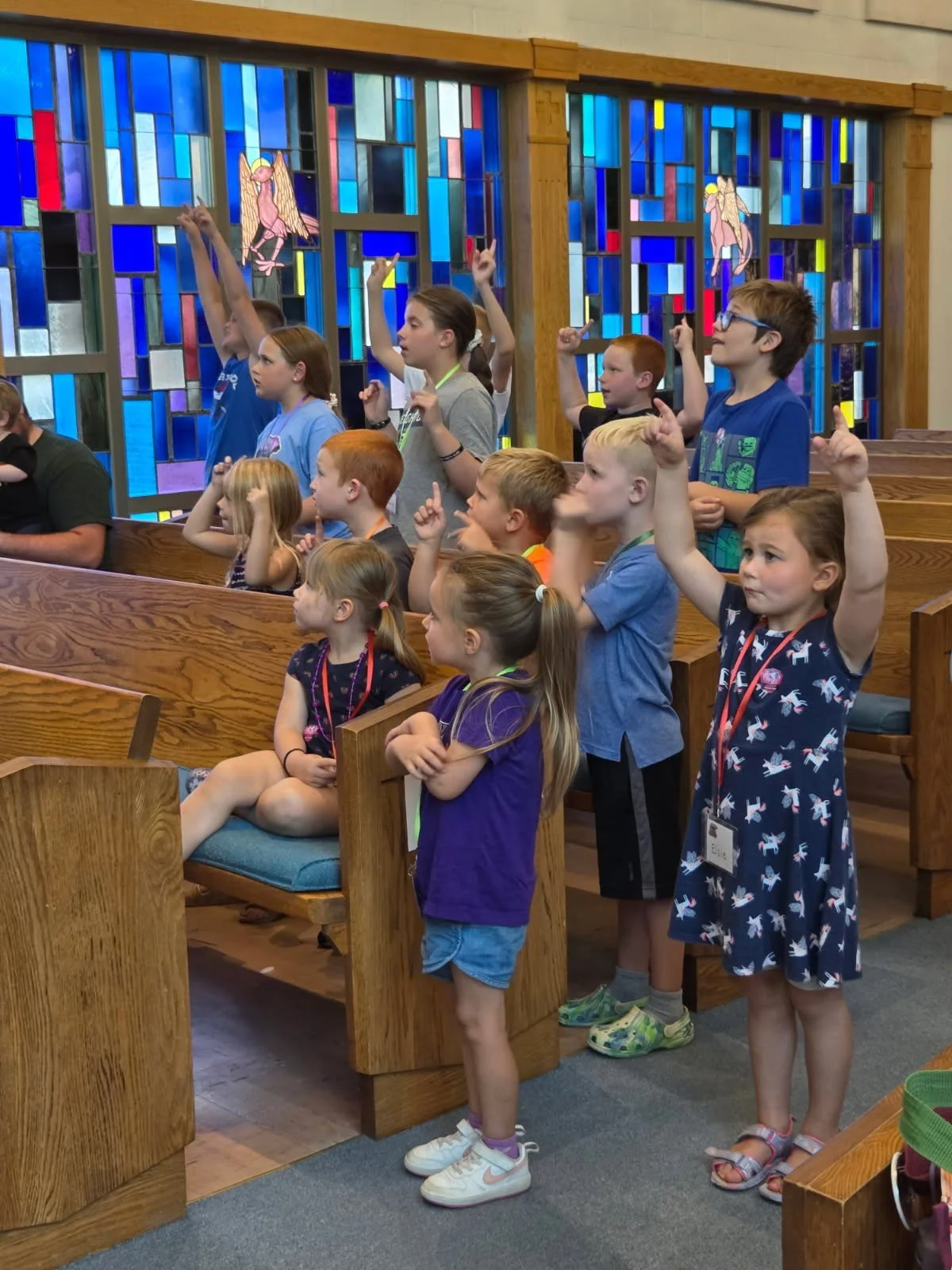 Group of children standing and sitting in a church, raising their hands, with colorful stained glass windows and religious artwork of angels in the background.