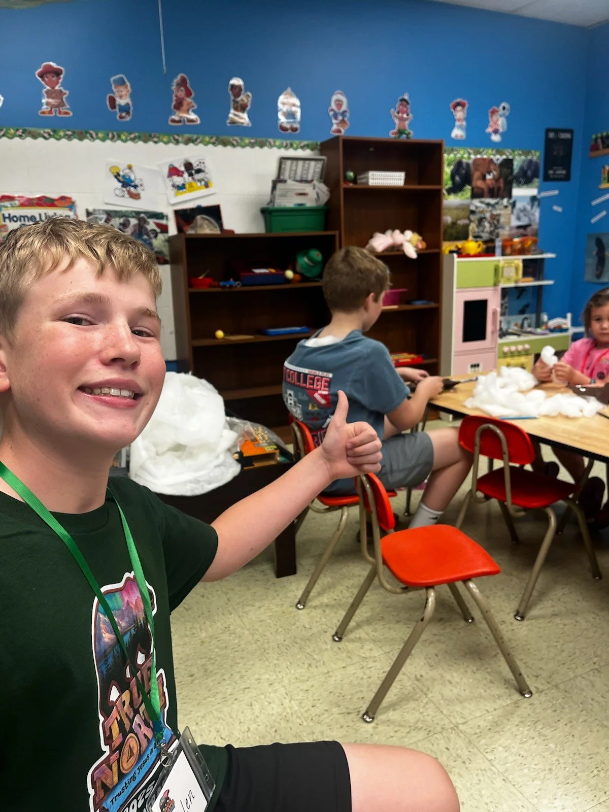 Smiling boy giving a thumbs up in a classroom with other children seated at a table, engaging in a craft activity involving cotton balls.