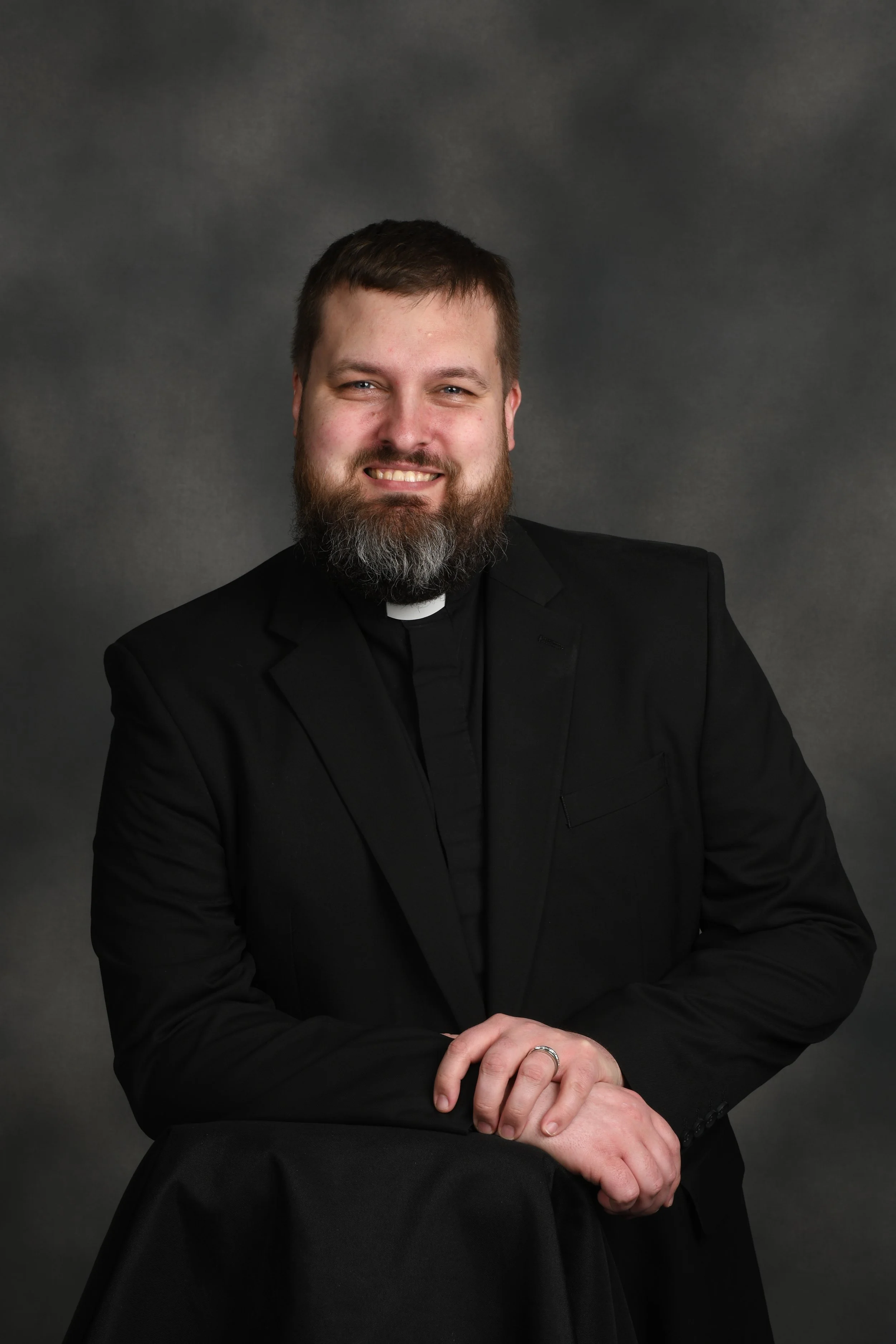 Portrait of a smiling man with a beard, wearing a black clerical shirt with a white collar and a black blazer, posed against a dark grey background.