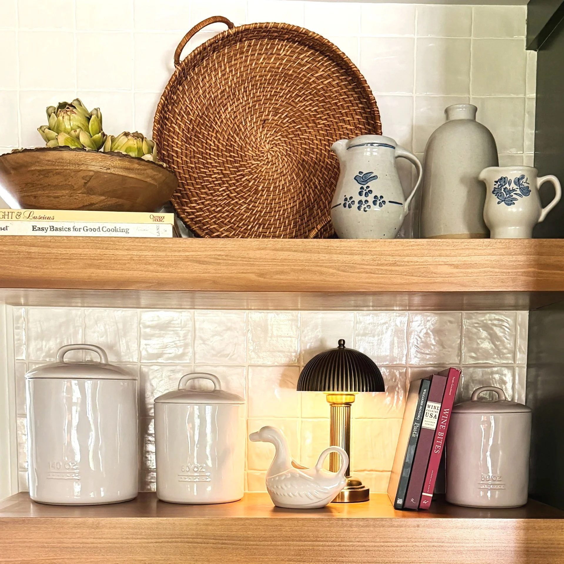 Decorative kitchen shelf with white ceramic containers, artichokes in a wooden bowl, woven tray, ceramic pitcher and creamer with blue patterns, black and gold table lamp, and cookbooks against a white tiled wall.