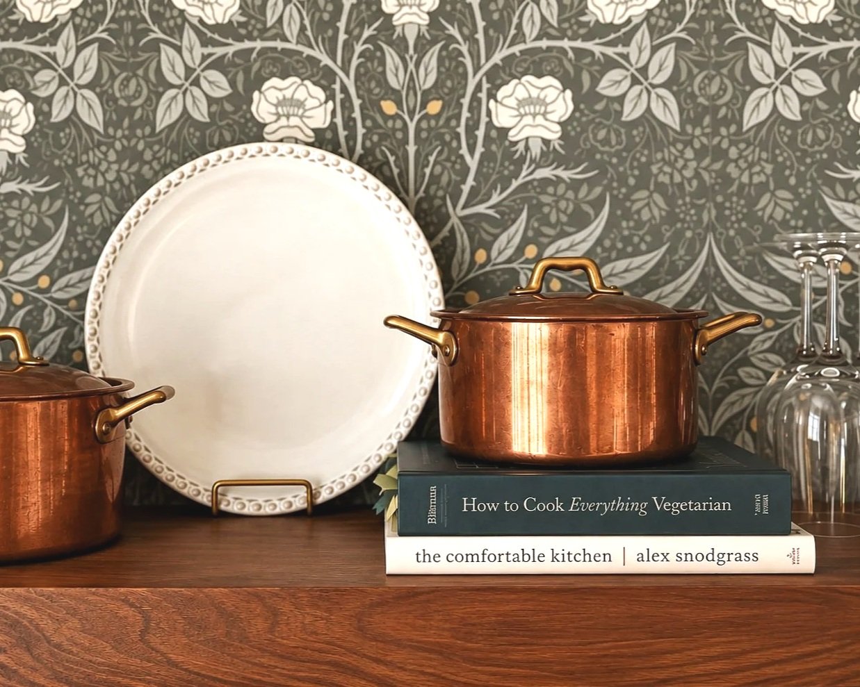 Copper pots, a white decorative plate, and two cookbooks on a wooden shelf in front of dark floral wallpaper.