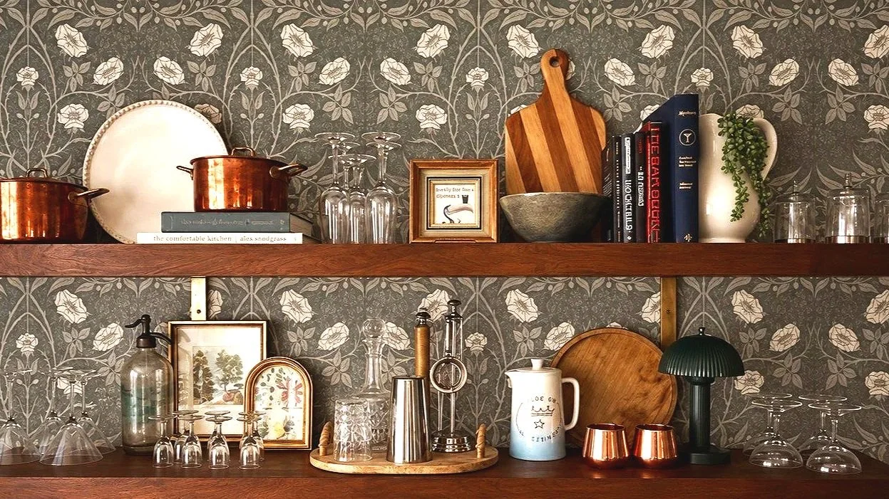 A decorated kitchen shelf with copper pots, glassware, framed pictures, books, cutting boards, a potted plant, and various decorative items against wallpaper with a floral pattern.
