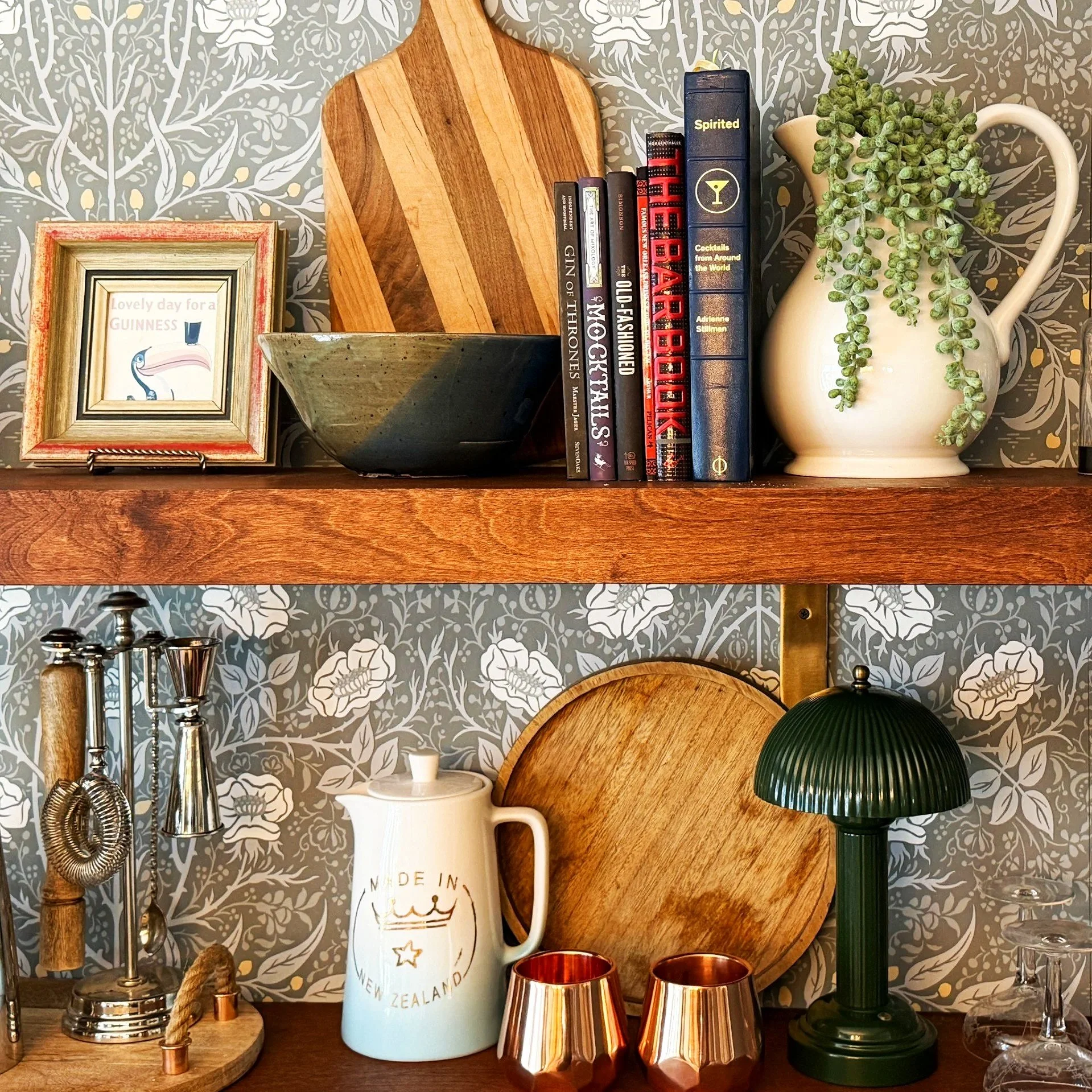 A shelf decorated with a picture frame, a wooden cutting board, a bowl, a row of books, a tall white pitcher with a green trailing plant, a green lamp, and copper cups, against a wallpaper with a floral pattern.