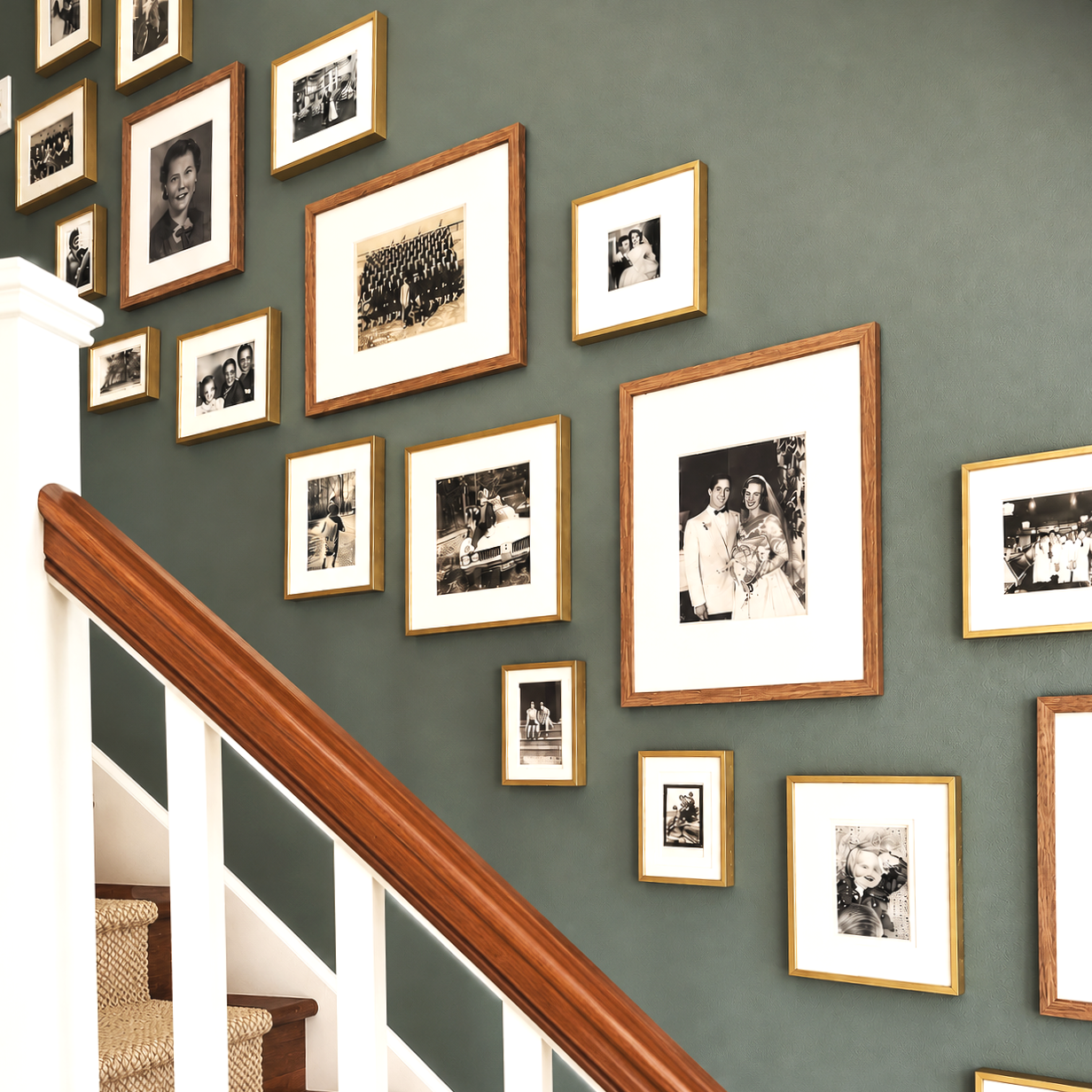 Wall decorated with a collection of black and white vintage photographs in gold and wood frames, positioned above a staircase with a wooden handrail and white spindles.