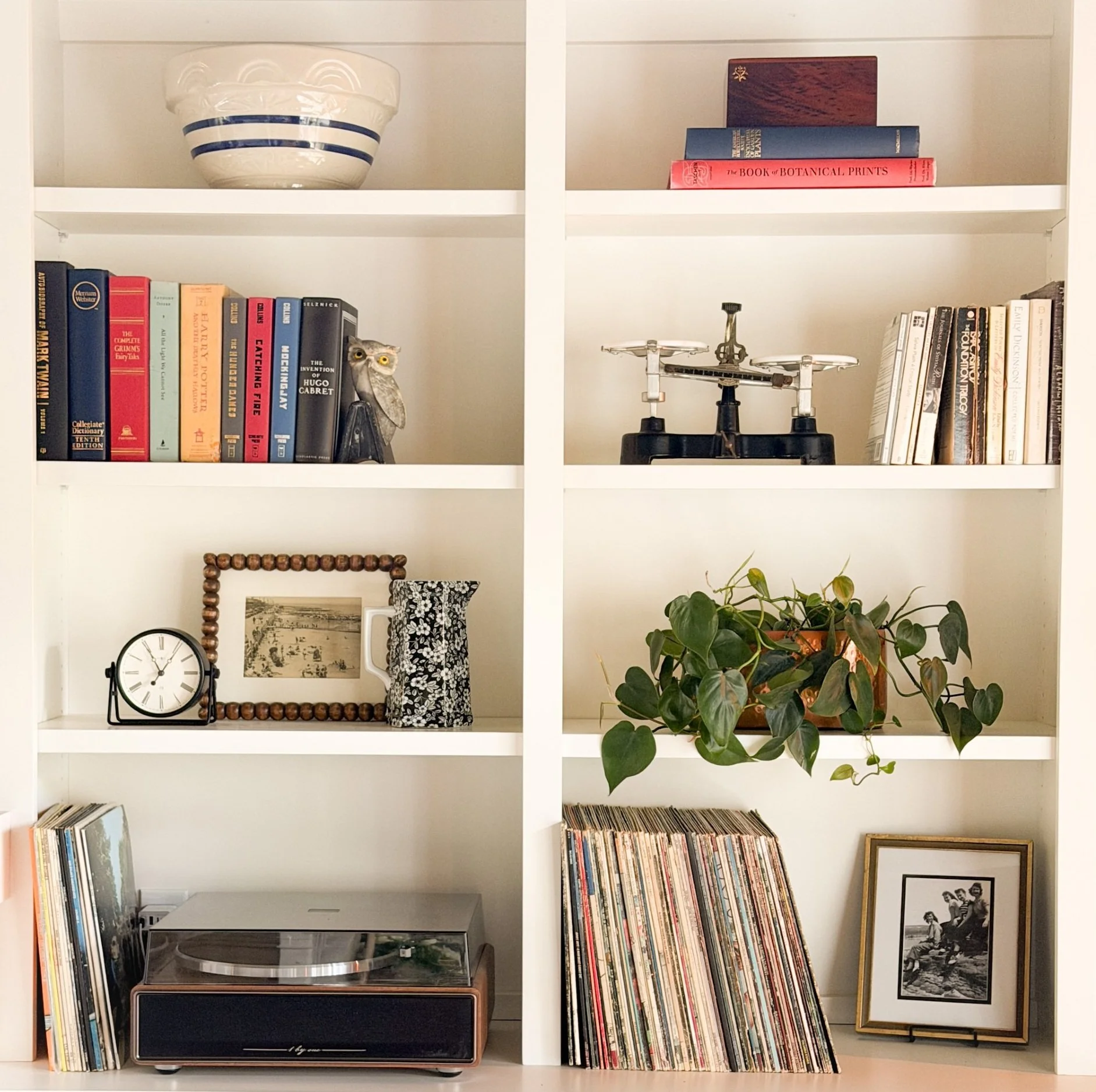White bookshelf with various books, decorative items, and a turntable. The top left contains a white bowl with blue stripes, the top middle has stacked books titled 'The Book of Botanical Prints'. The middle shelves have more books, a owl figurine, a