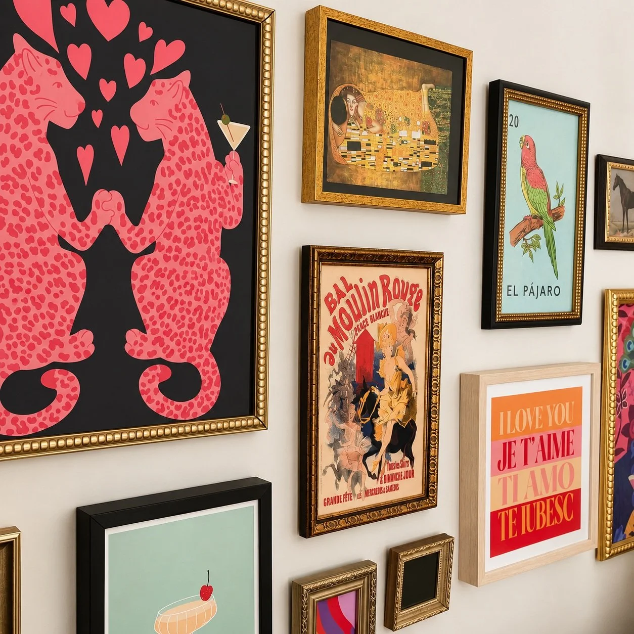 A gallery wall featuring various framed art and posters, including illustrations of cats with hearts, a classical painting, a poster for Moulin Rouge, a colorful bird labeled "El Pájaro," a "I Love You" message in multiple languages, and other decora