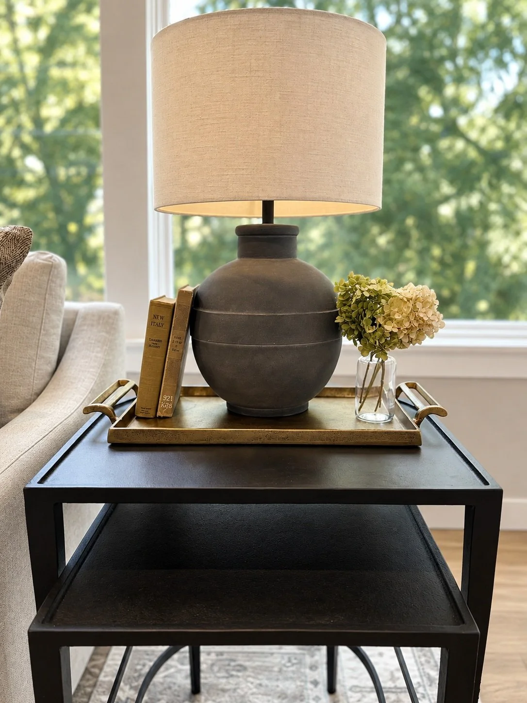 A black side table with a gold tray on top, holding a large gray ceramic lamp with a beige lampshade, a small glass vase with white and green hydrangea flowers, and two beige books, with a beige sofa and window with green trees in the background.