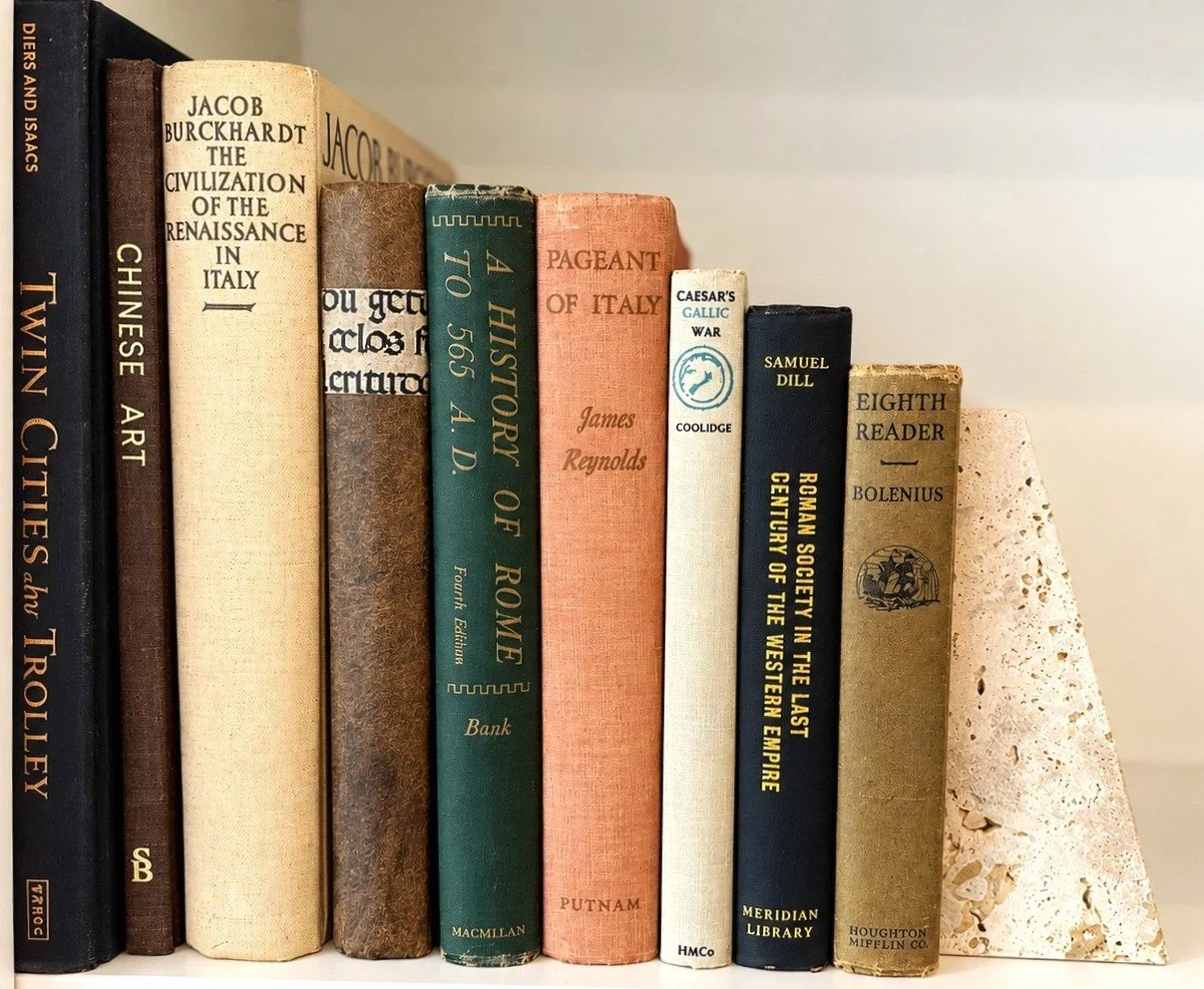 A collection of hardcover books arranged vertically on a shelf, with titles and authors visible on their spines, including a decorative piece resembling a triangular sculpture made of porous material on the right.