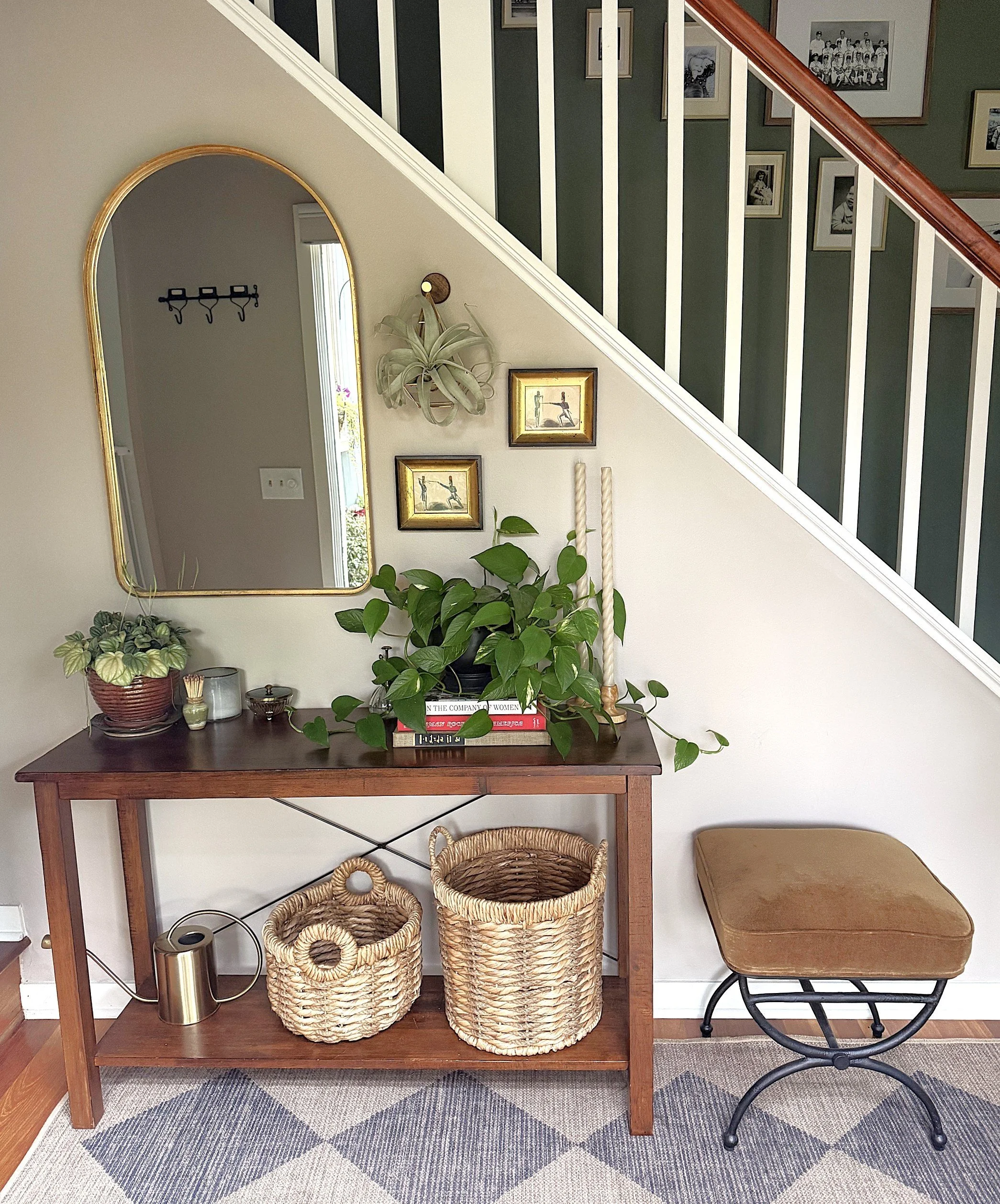 A hallway with a wooden console table decorated with plants, candles, and books. Above the table hangs a large mirror with a gold frame. Baskets are placed on the floor under the table. To the right, there is a small upholstered stool with a metal ba