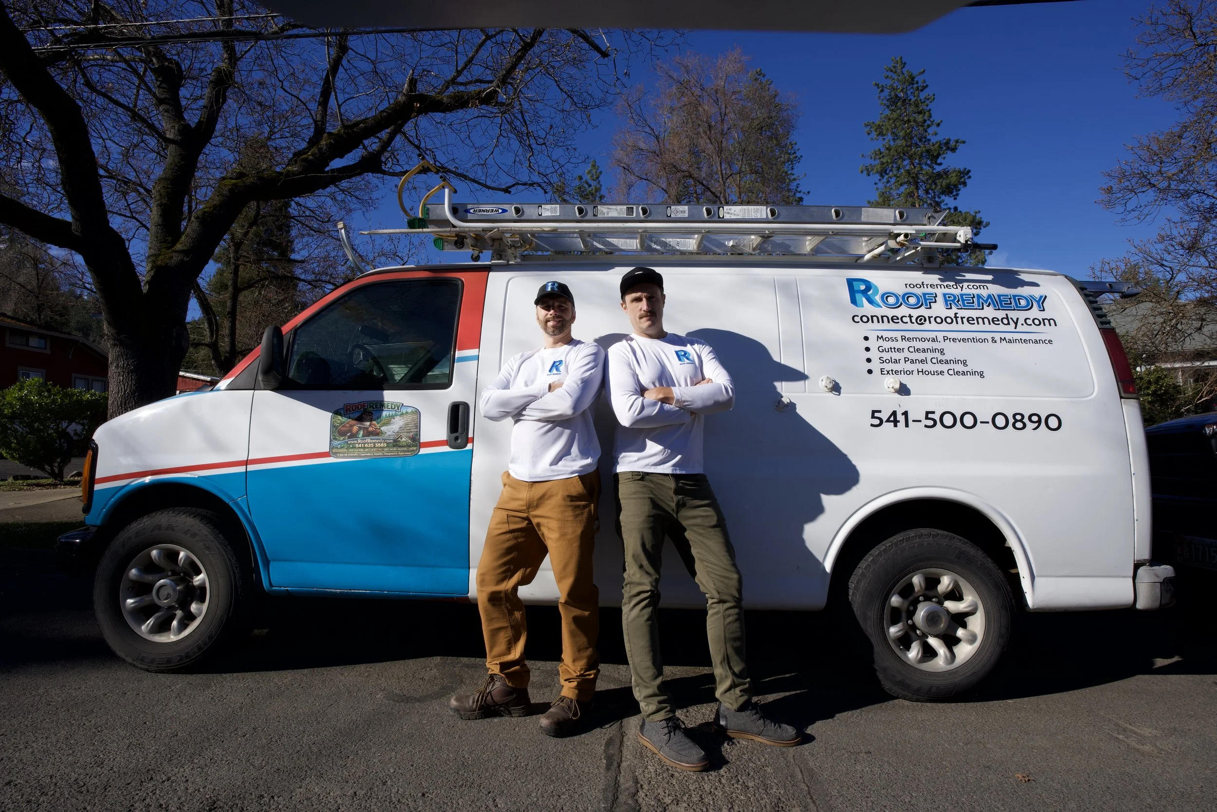 Two men standing with arms crossed in front of a company van with roofing services advertisements. The van is white with blue and red accents, has a ladder on top, and displays roofing service details including contact information. The men are wearing white long-sleeve shirts with company logos, one with brown pants and the other with green pants. They are outdoors on a sunny day with trees and houses in the background.
