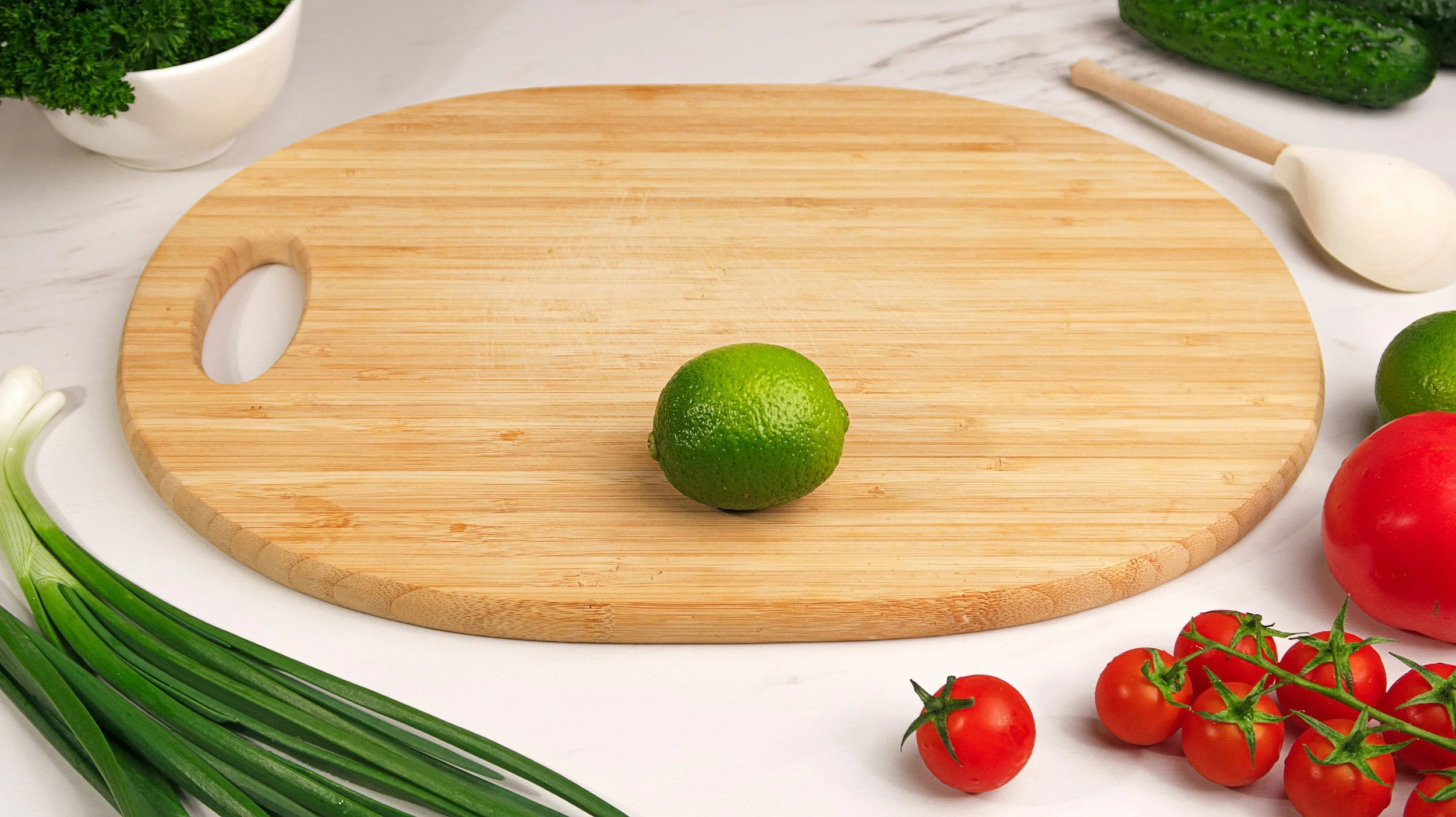 A round bamboo cutting board on a white countertop, with a lime in the center. Surrounding the board are cherry tomatoes on a vine, a cucumber, green onions, a bunch of parsley, and a bowl of parsley.
