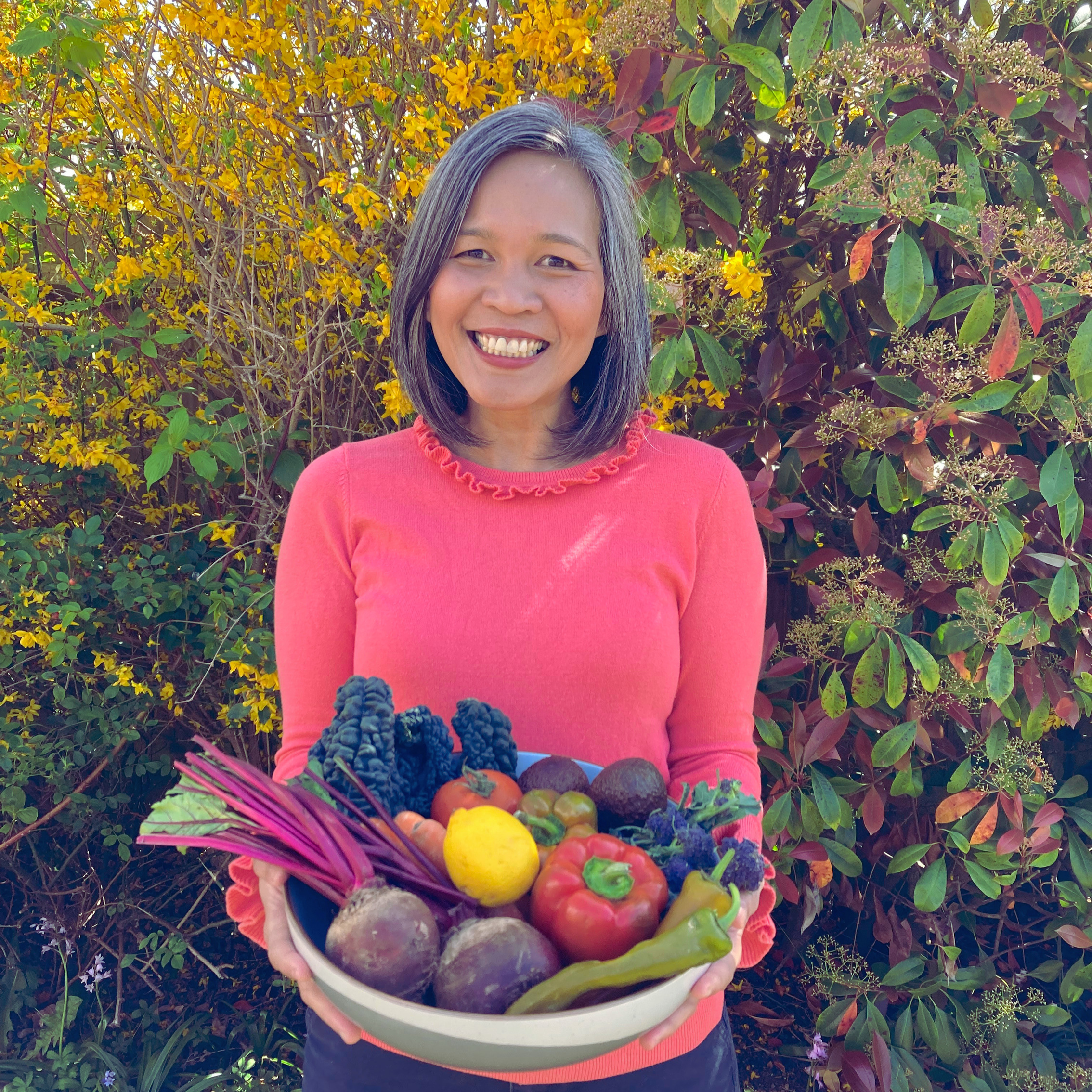 A woman smiling outdoors holding a large bowl of fresh vegetables and fruits. The background features yellow and green foliage.