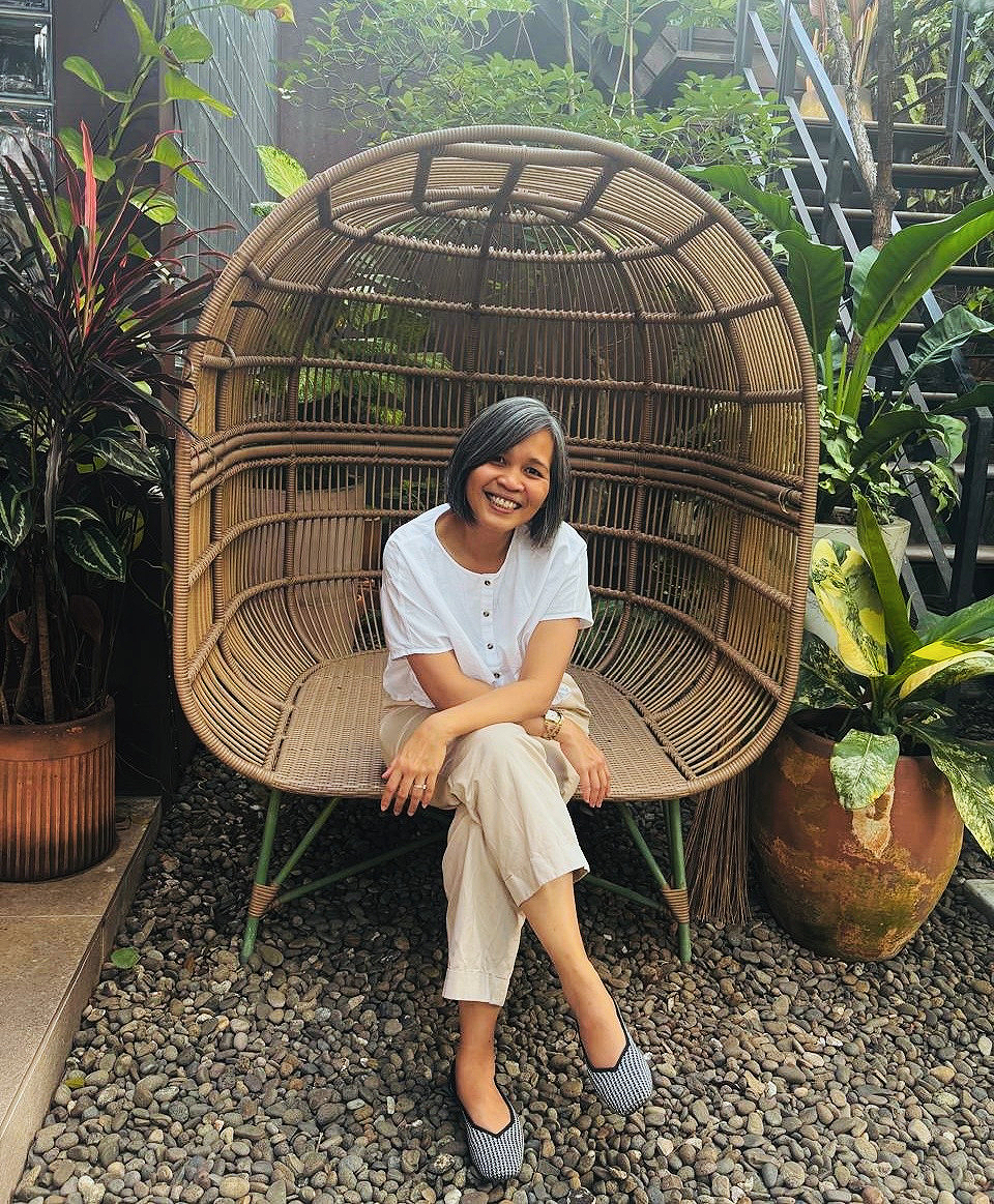A woman smiling sitting on a rattan outdoor chair surrounded by green plants and potted foliage.