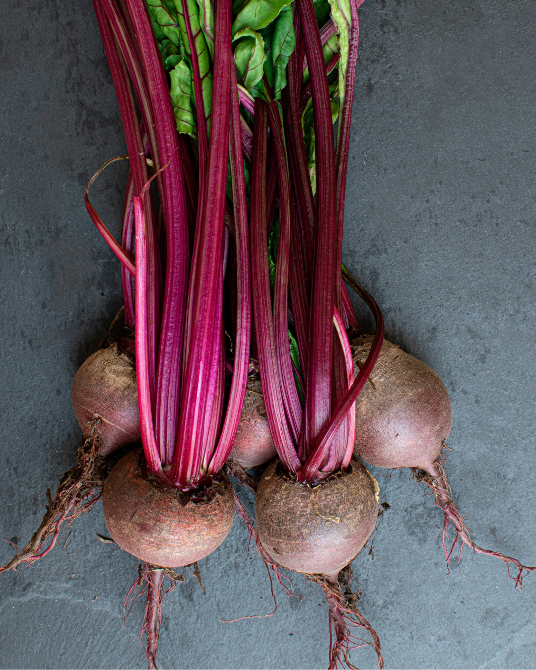 Fresh beets with green leaves and roots on a dark surface.