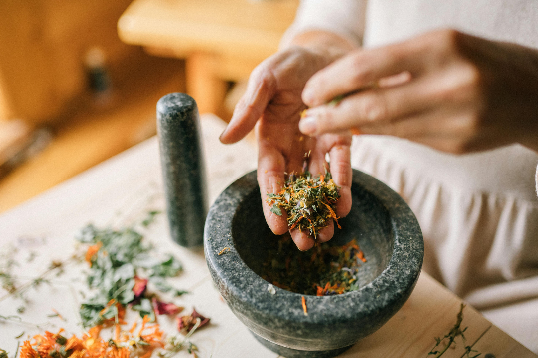 Person grinding dried herbs in a mortar and pestle with loose herbs and flowers on a wooden table.