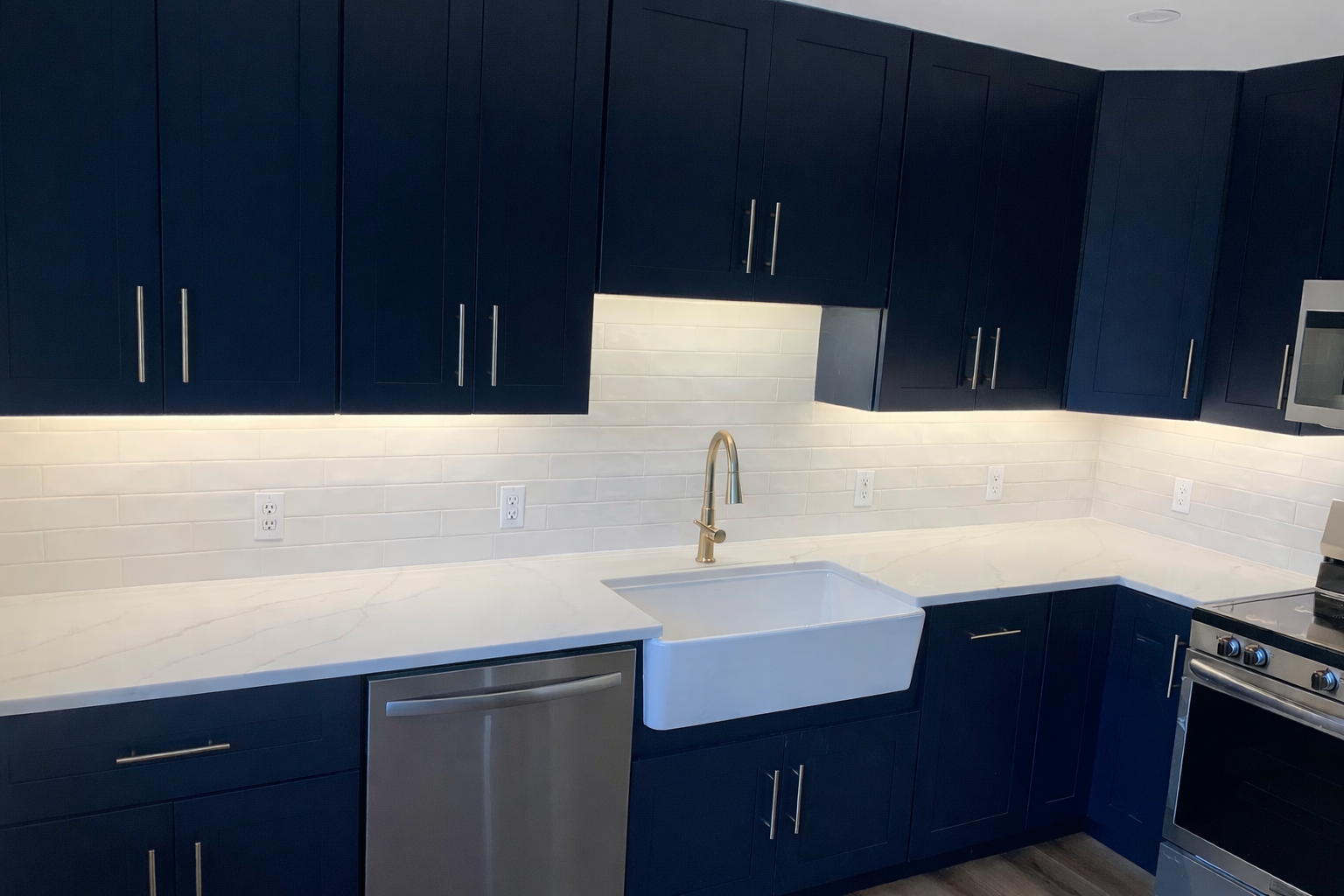 Modern kitchen with navy blue cabinets, marble countertop, white farmhouse sink, stainless steel dishwasher, and stove, white subway tile backsplash, and multiple electrical outlets.