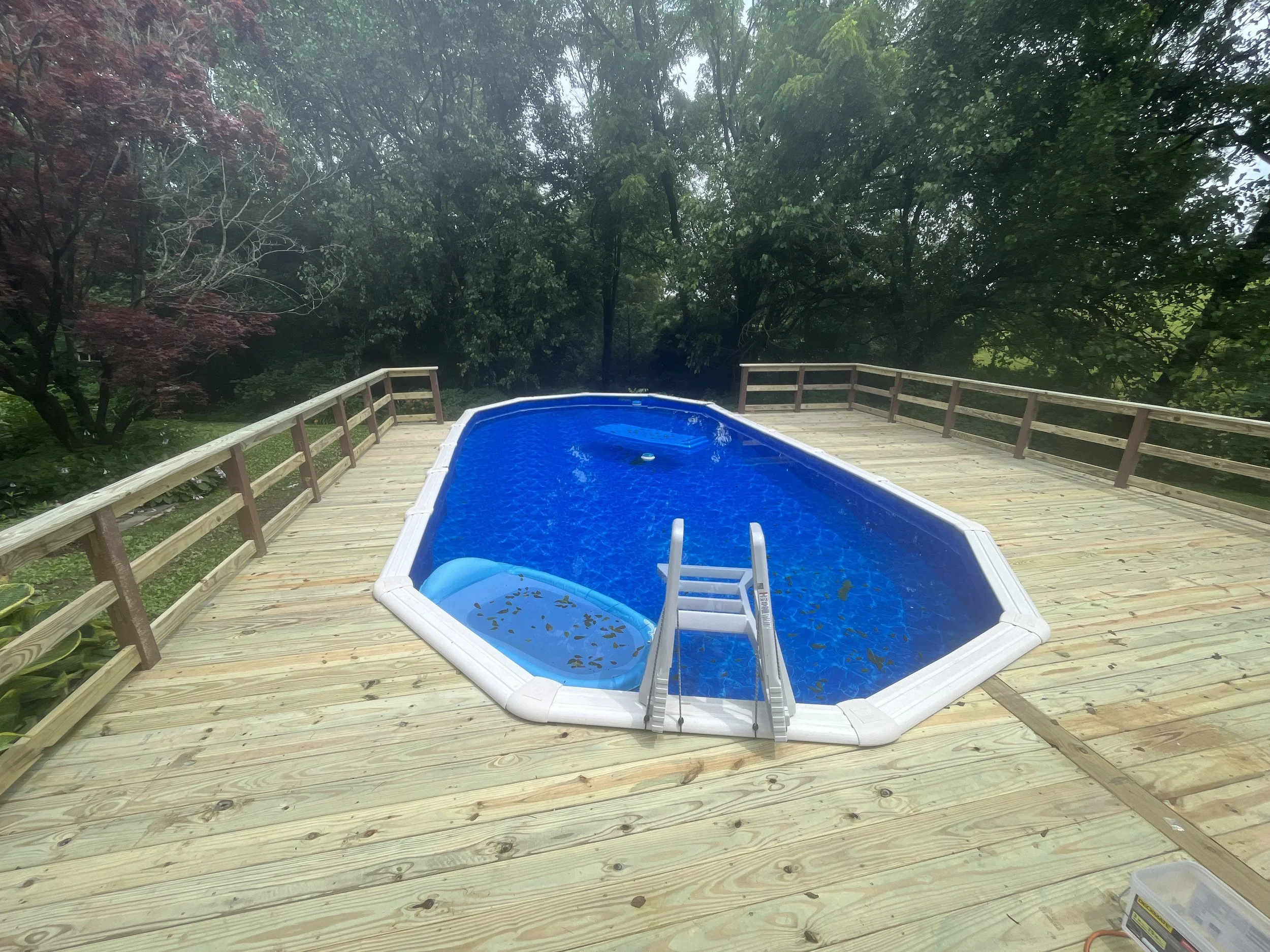 An outdoor above-ground swimming pool on a wooden deck surrounded by trees, with leaves floating in the water and a pool ladder.