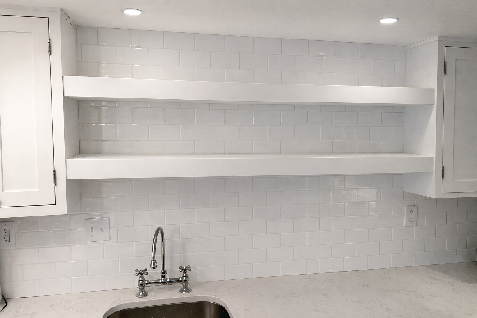 Empty white kitchen shelves above a countertop and faucet, with white tiled backsplash and cabinet doors on either side.