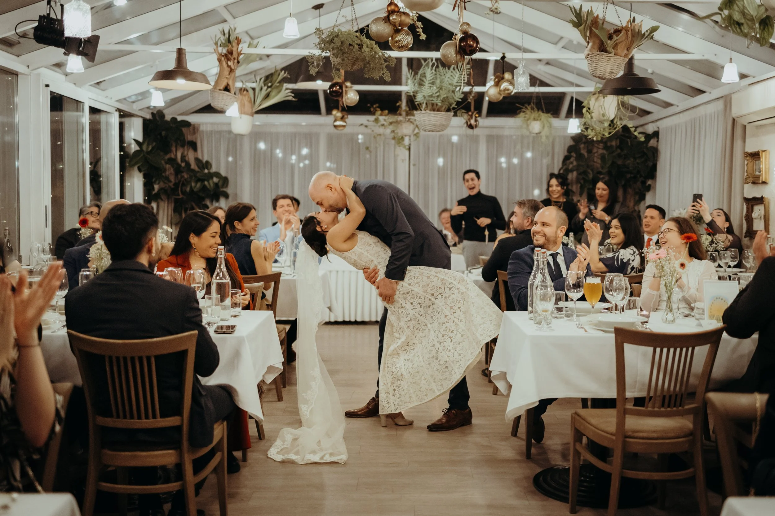 A bride and groom dance in the center of a wedding reception hall, surrounded by seated guests clapping and smiling.