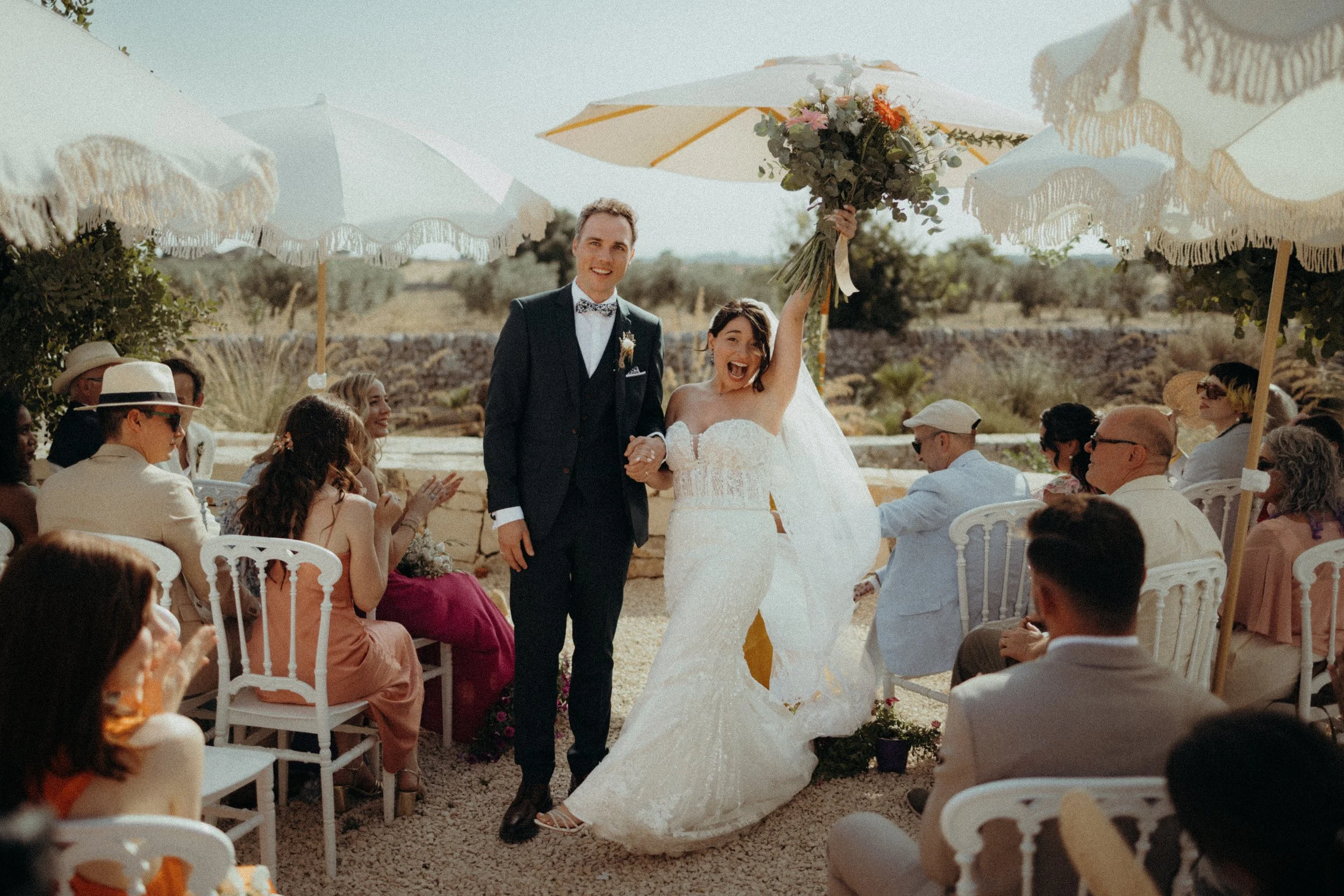 A newlywed couple walking down the aisle at their outdoor wedding, surrounded by seated guests under umbrellas on a sunny day.