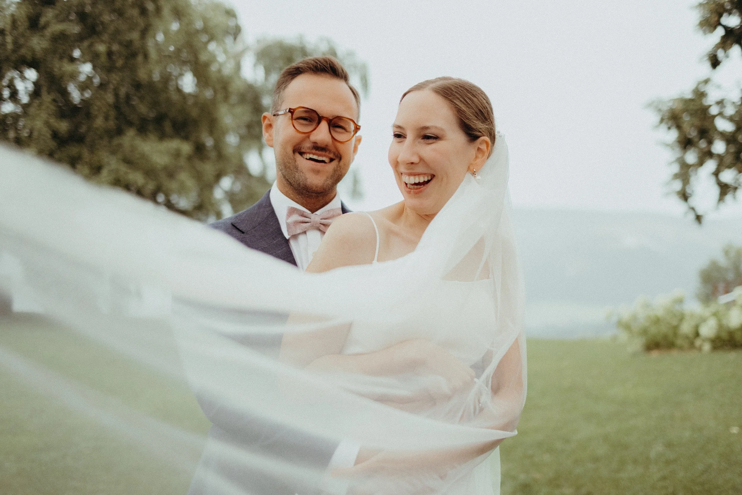 A happy bride and groom smiling outdoors, with the bride's veil flowing in the foreground and trees in the background.