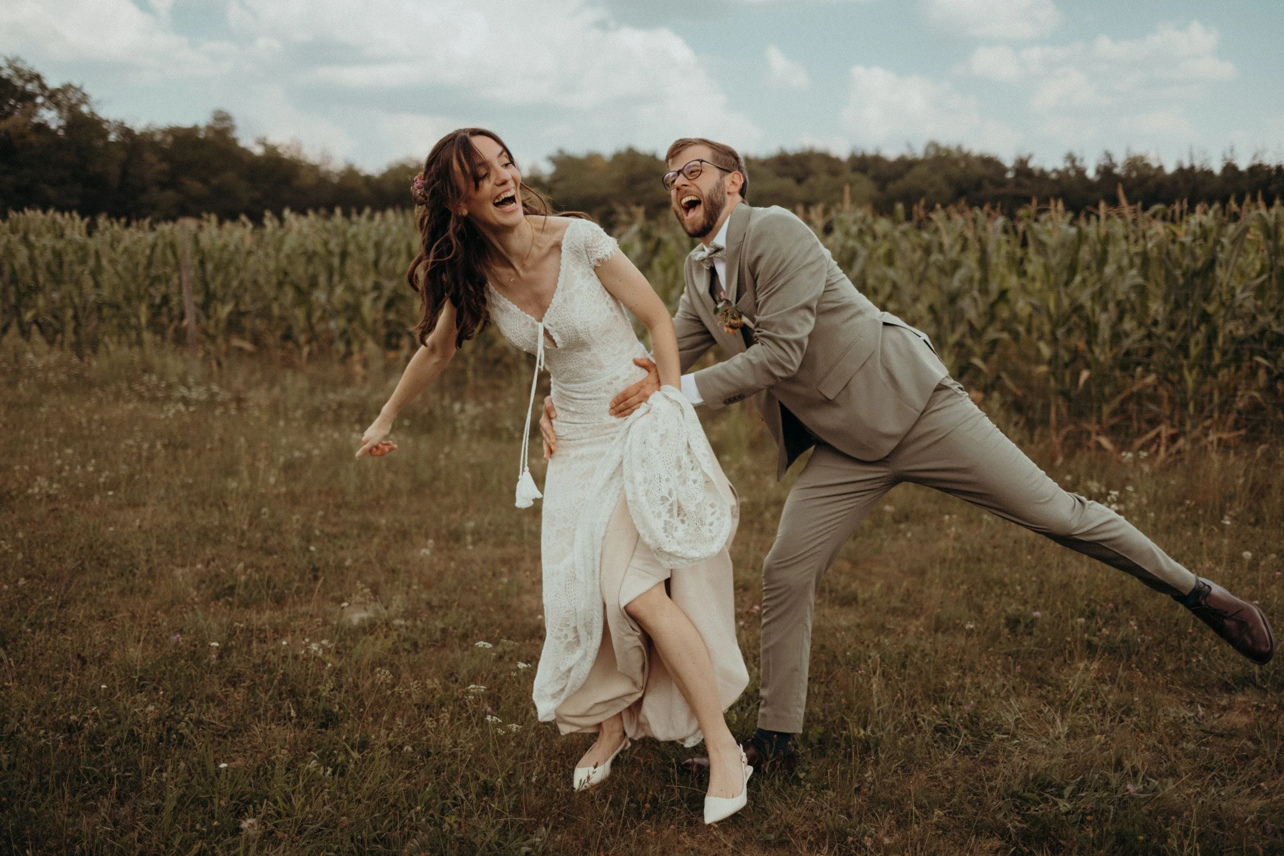 A joyful couple dancing in a field with a cornfield in the background. The woman wears a white lace wedding dress, and the man is in a light gray suit with glasses. Both are smiling and laughing.