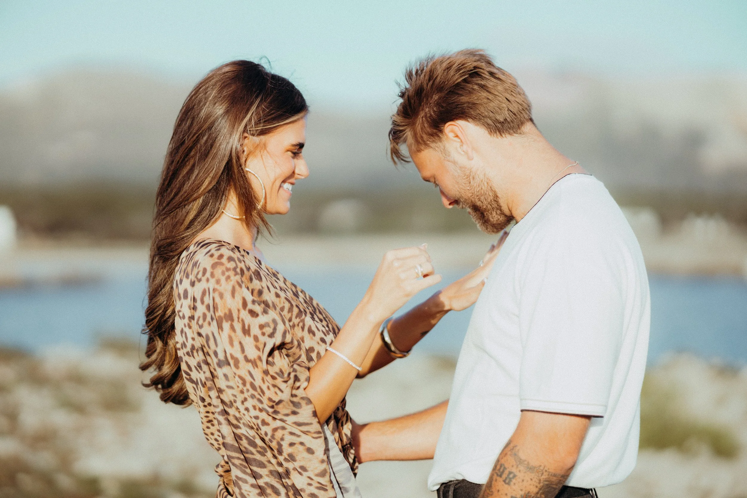 A woman in a leopard print top and a man in a white shirt are standing outdoors near water, smiling and touching at the chest during a romantic moment.