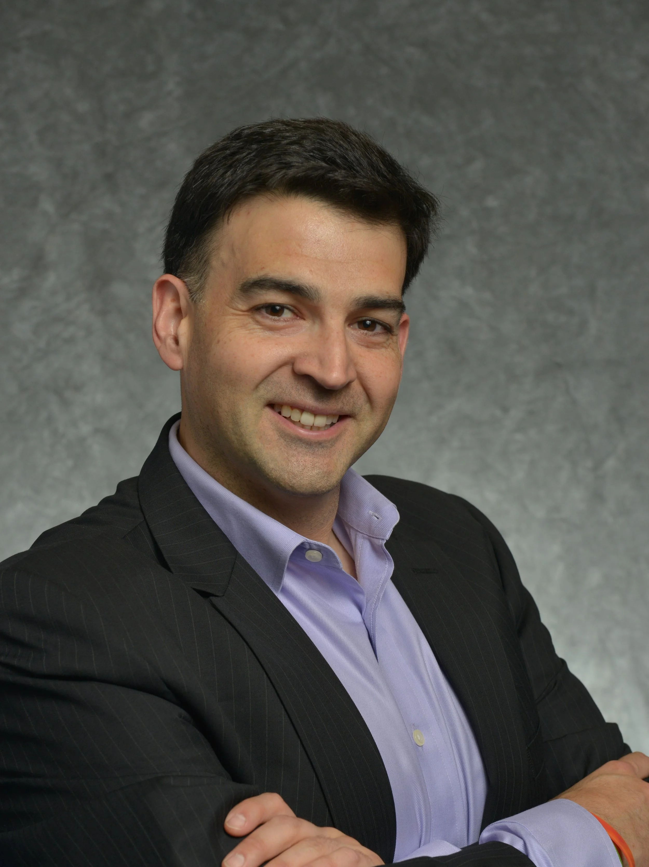 A man with dark hair and a light purple shirt, wearing a dark pinstripe suit, smiling against a grey textured background.
