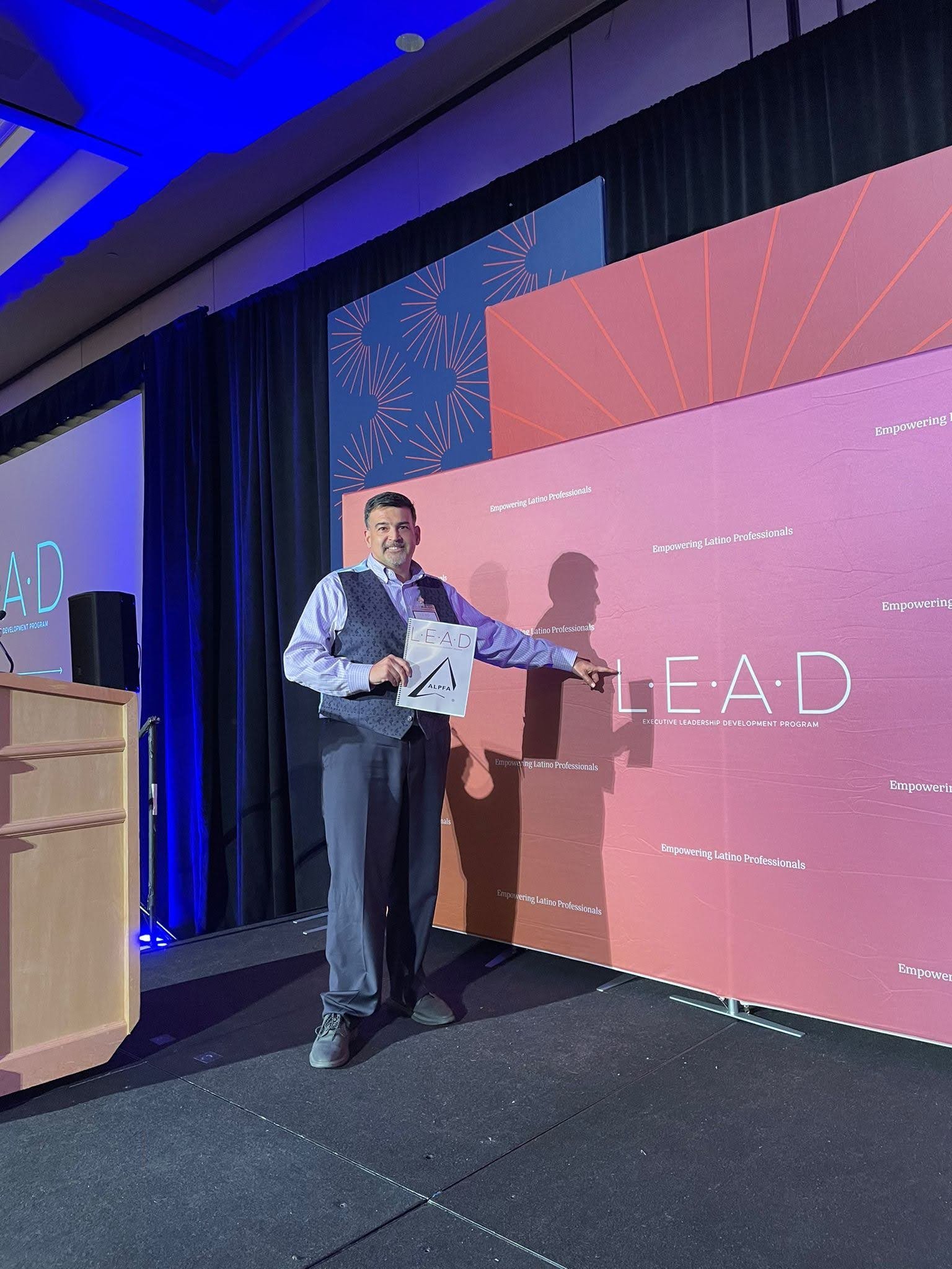 Eduardo Villavicencio holding a "L.E.A.D." sign on stage at an Executive Leadership Development Program event for Latino professionals