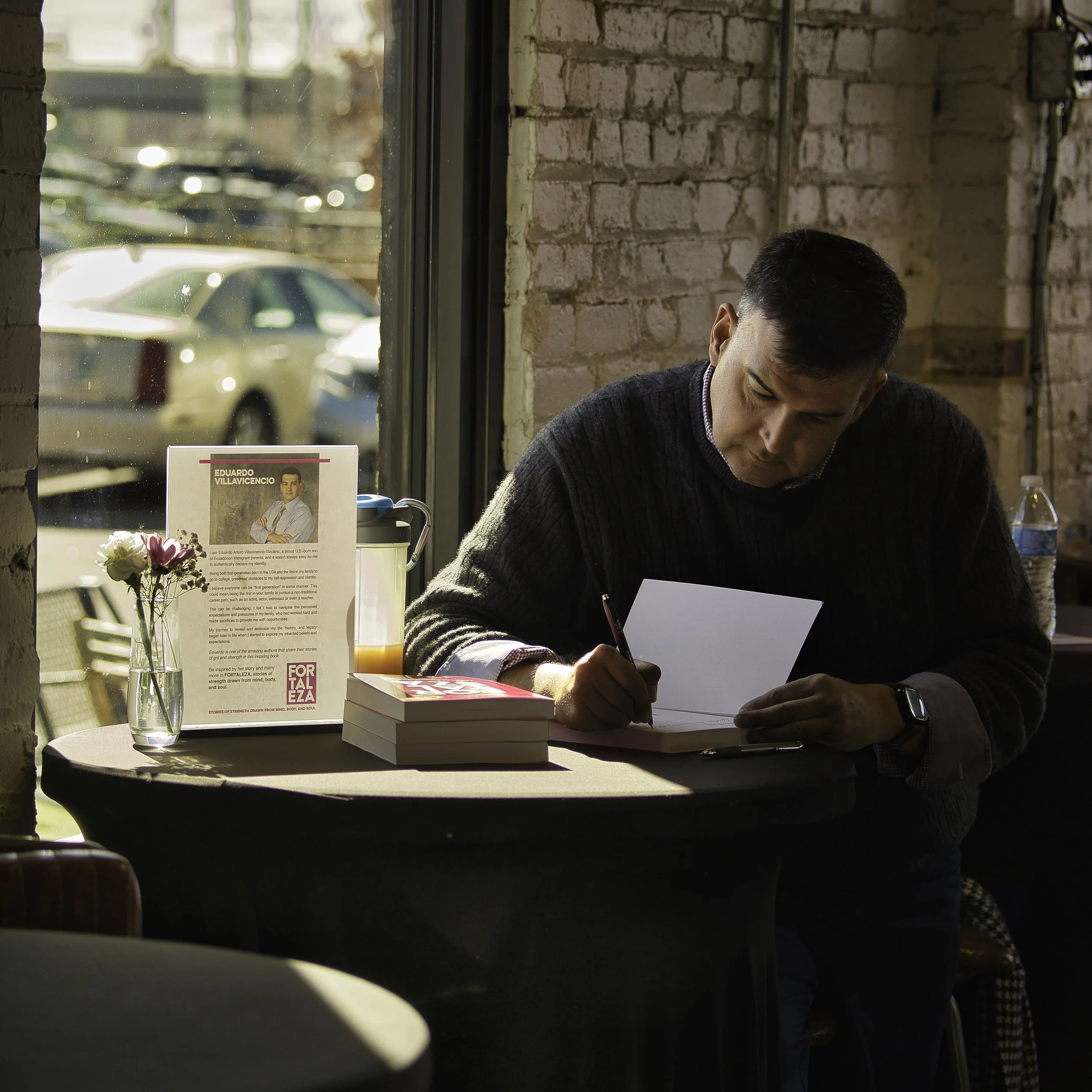 Eduardo Villavicencio writing at a table, with his book Fortaleza displayed alongside him