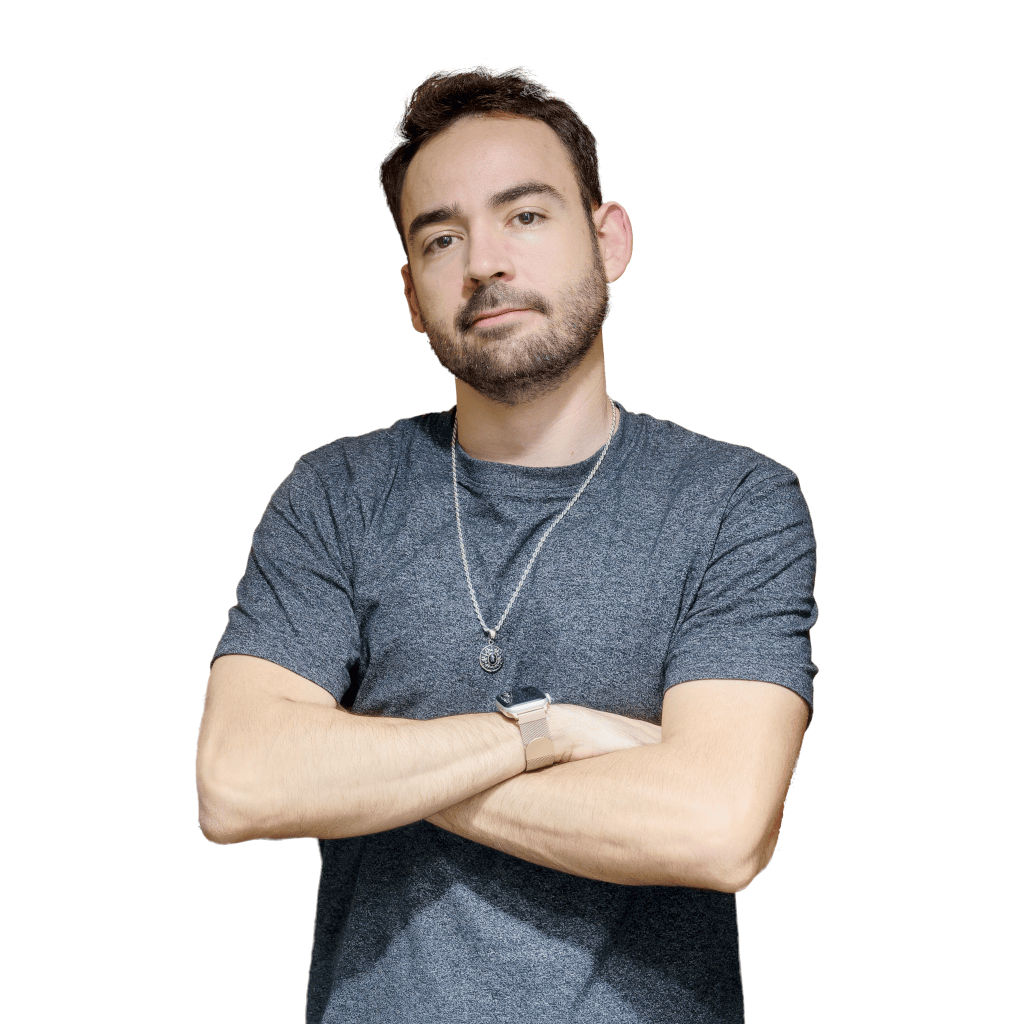 A young man with dark hair, beard, and mustache, wearing a gray t-shirt, a necklace, a wristwatch, and crossed arms, looking confidently at the camera.