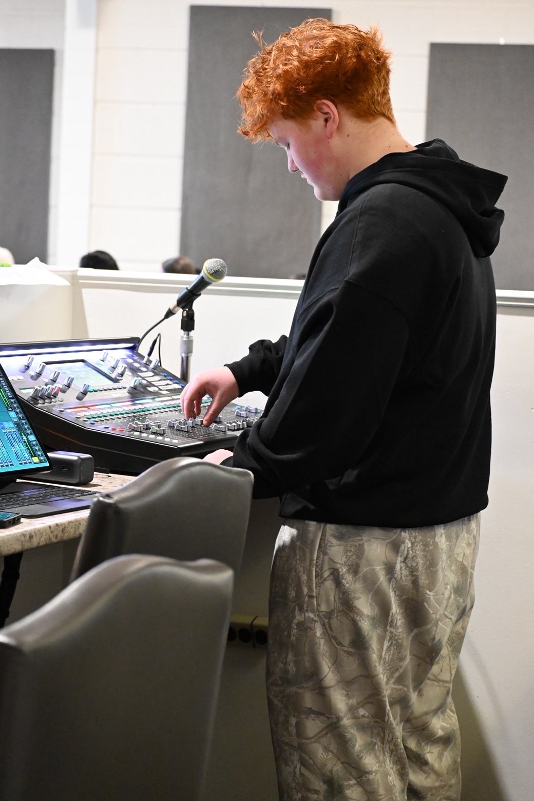 A young man with curly red hair wearing a black hoodie and camouflage pants working on a sound mixing console at a table, with a microphone nearby.