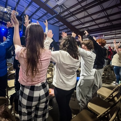 People standing in a row with hands raised during a worship event inside a large indoor venue with a high ceiling.