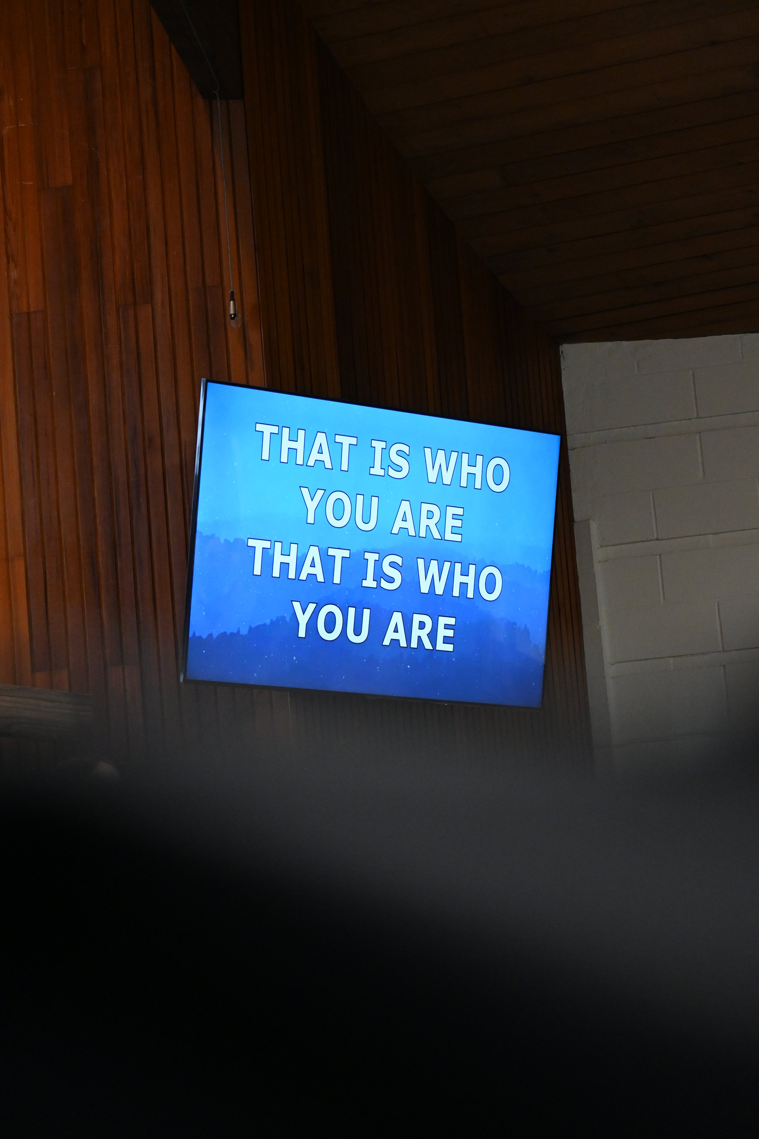 A digital screen displaying the lyrics to the worship song "Way Maker." The lyrics read 'That is who you are, that is who you are' against a backdrop of blue mountains and trees, mounted on a wooden wall.
