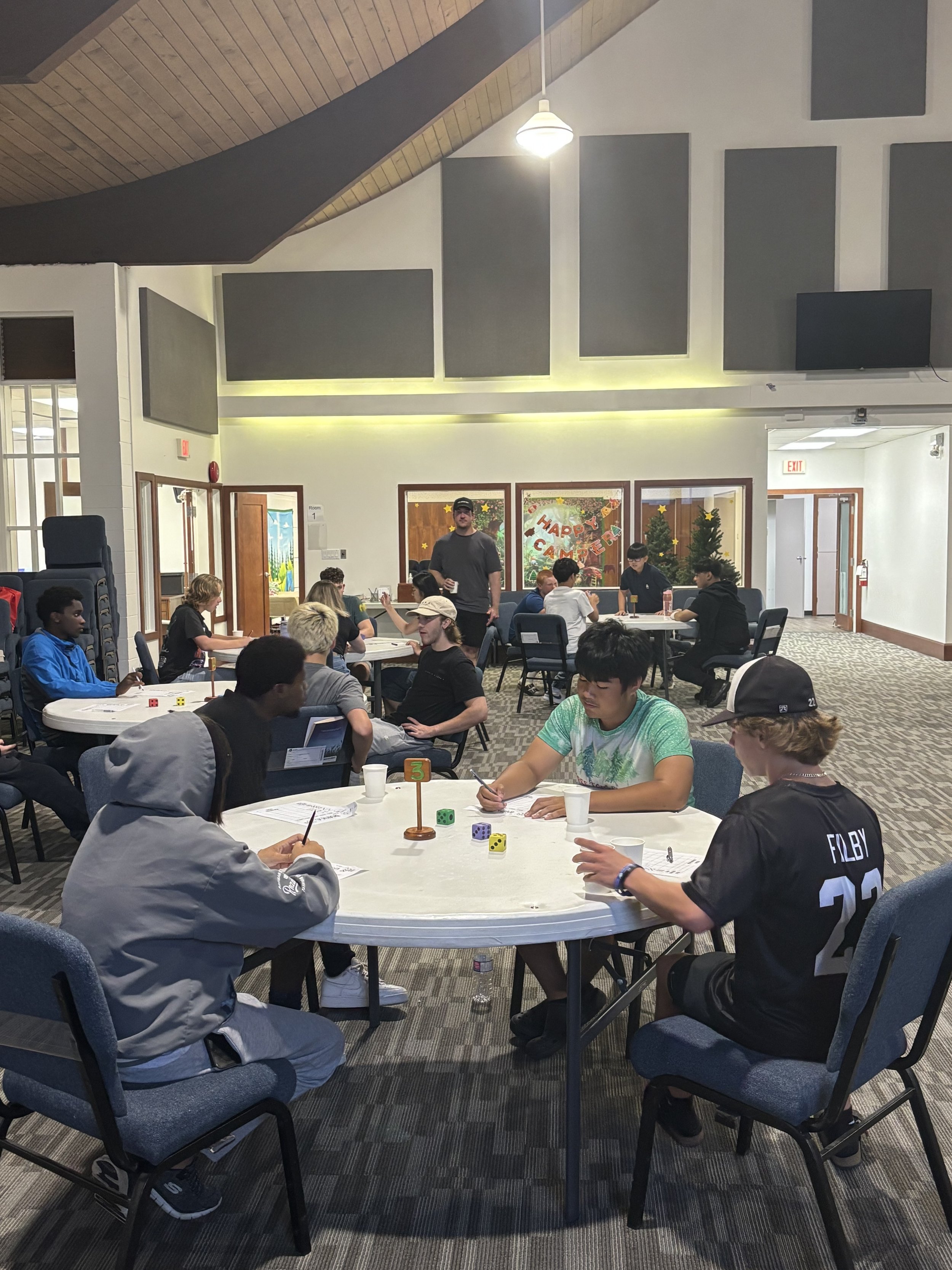 Young people sitting around tables playing a board game at a youth event in a church, with some attendees engaged in conversation and others rolling dice.