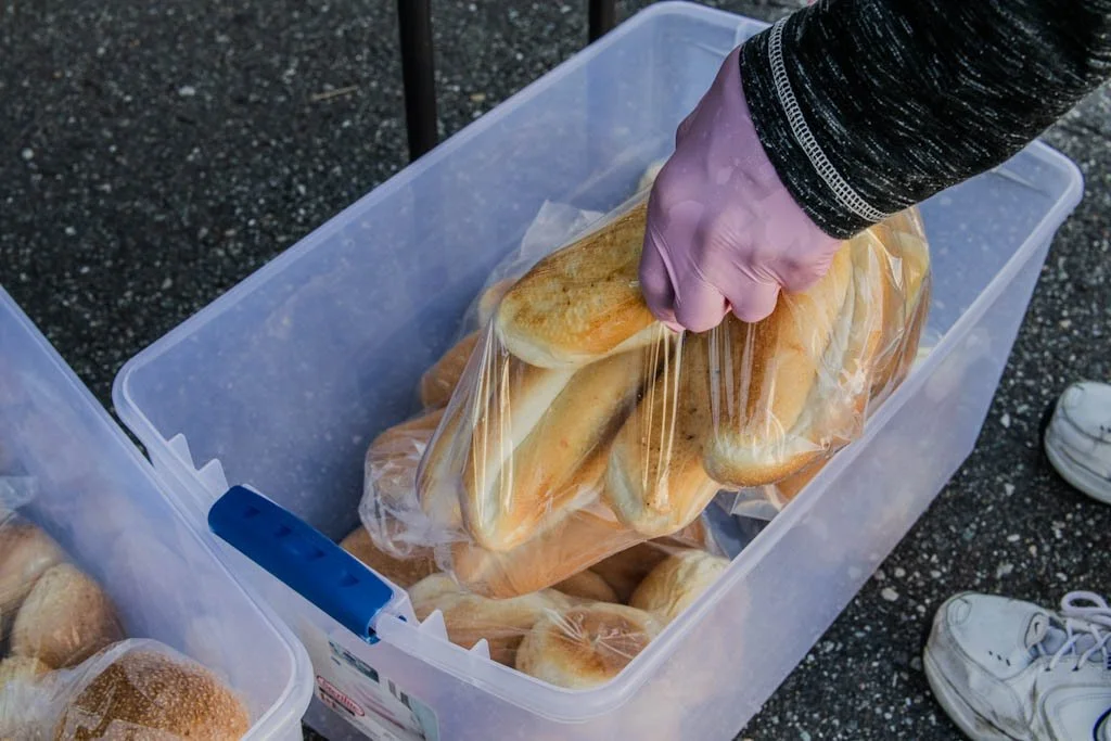 Person wearing gloves placing bread rolls into a clear plastic bag inside a large plastic container on a gravel surface.