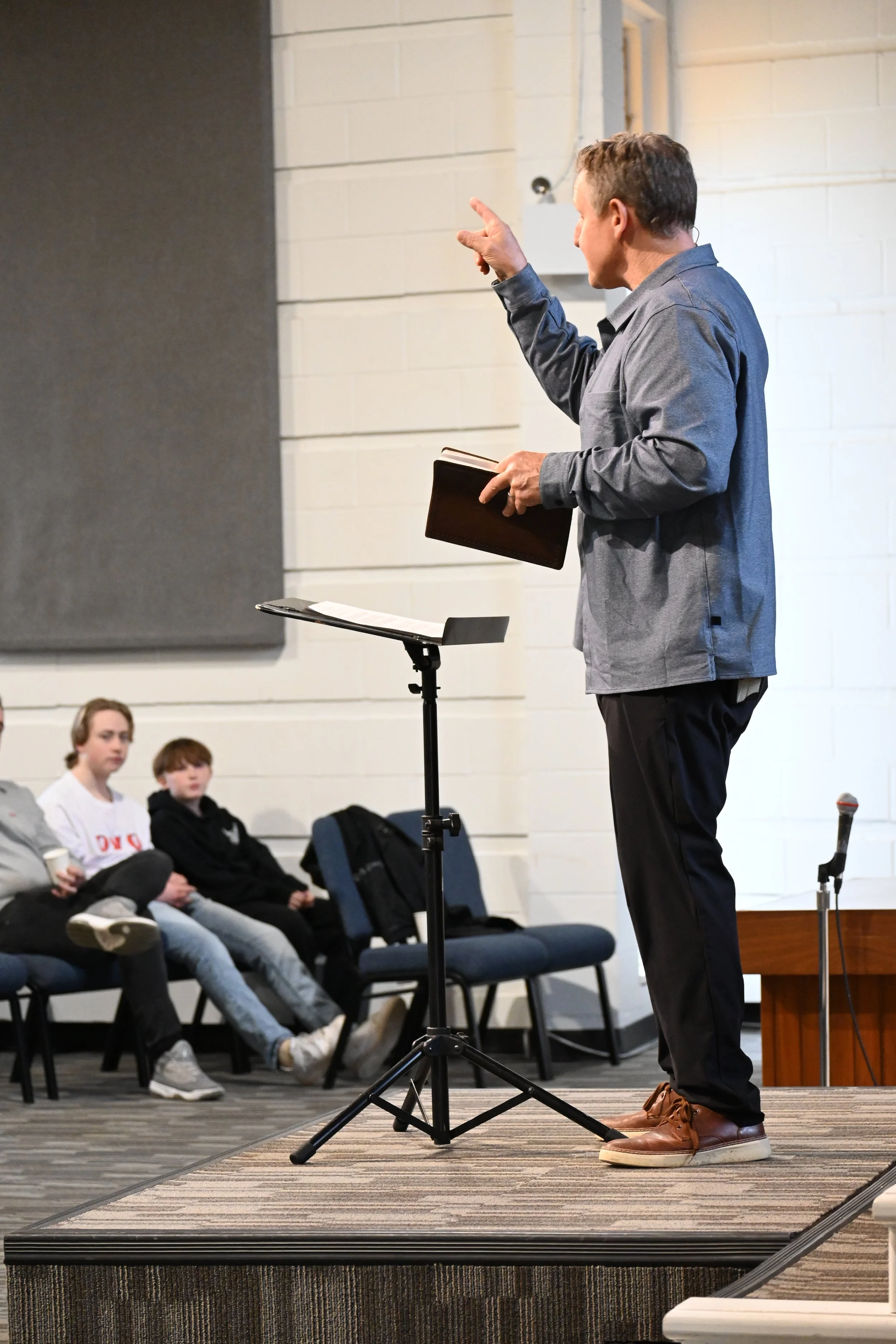 A man giving a speech or lecture on stage with a music stand and microphone, with children sitting in the audience watching.