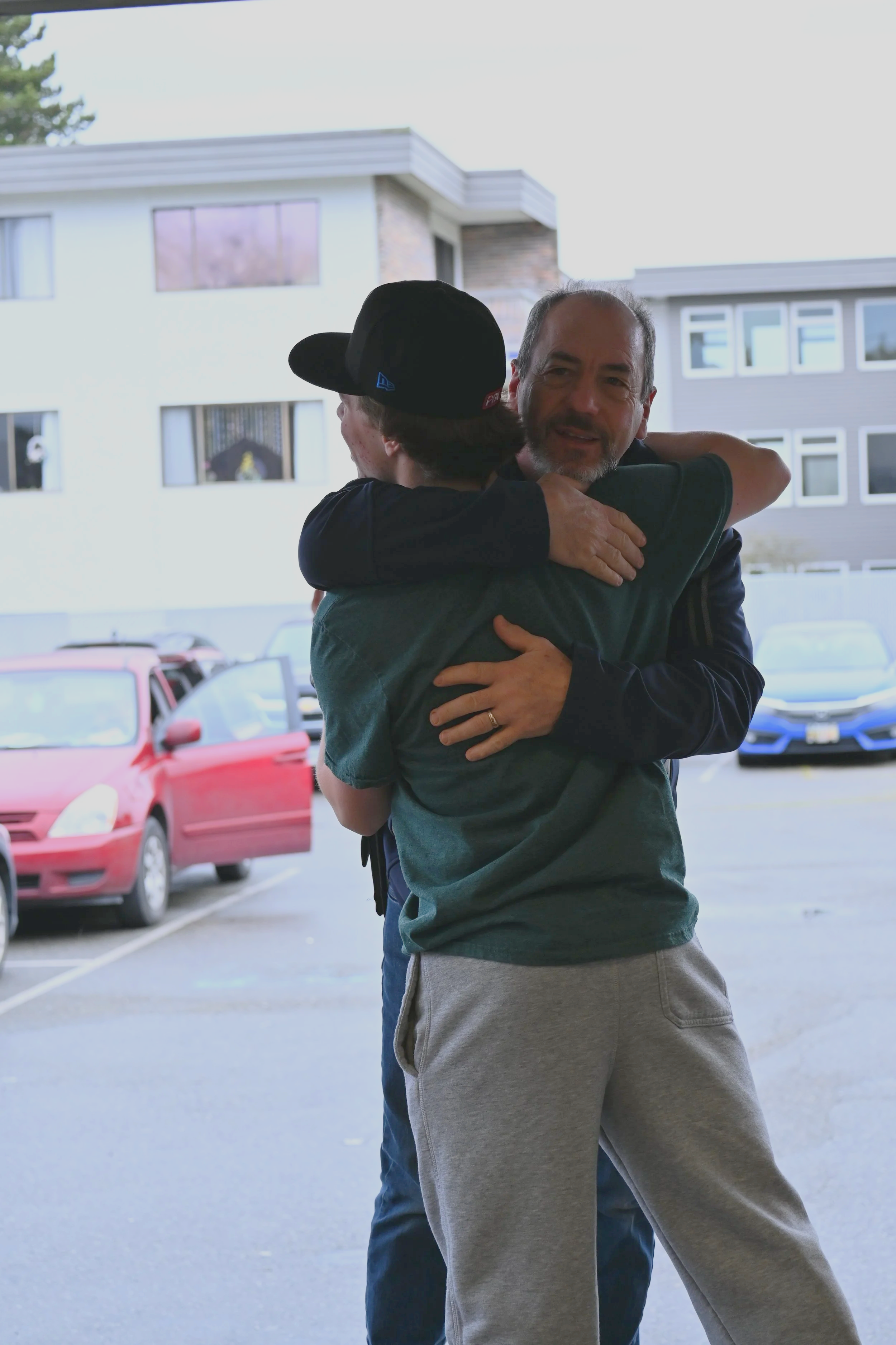 Two men hugging outside near parked cars with buildings in the background.