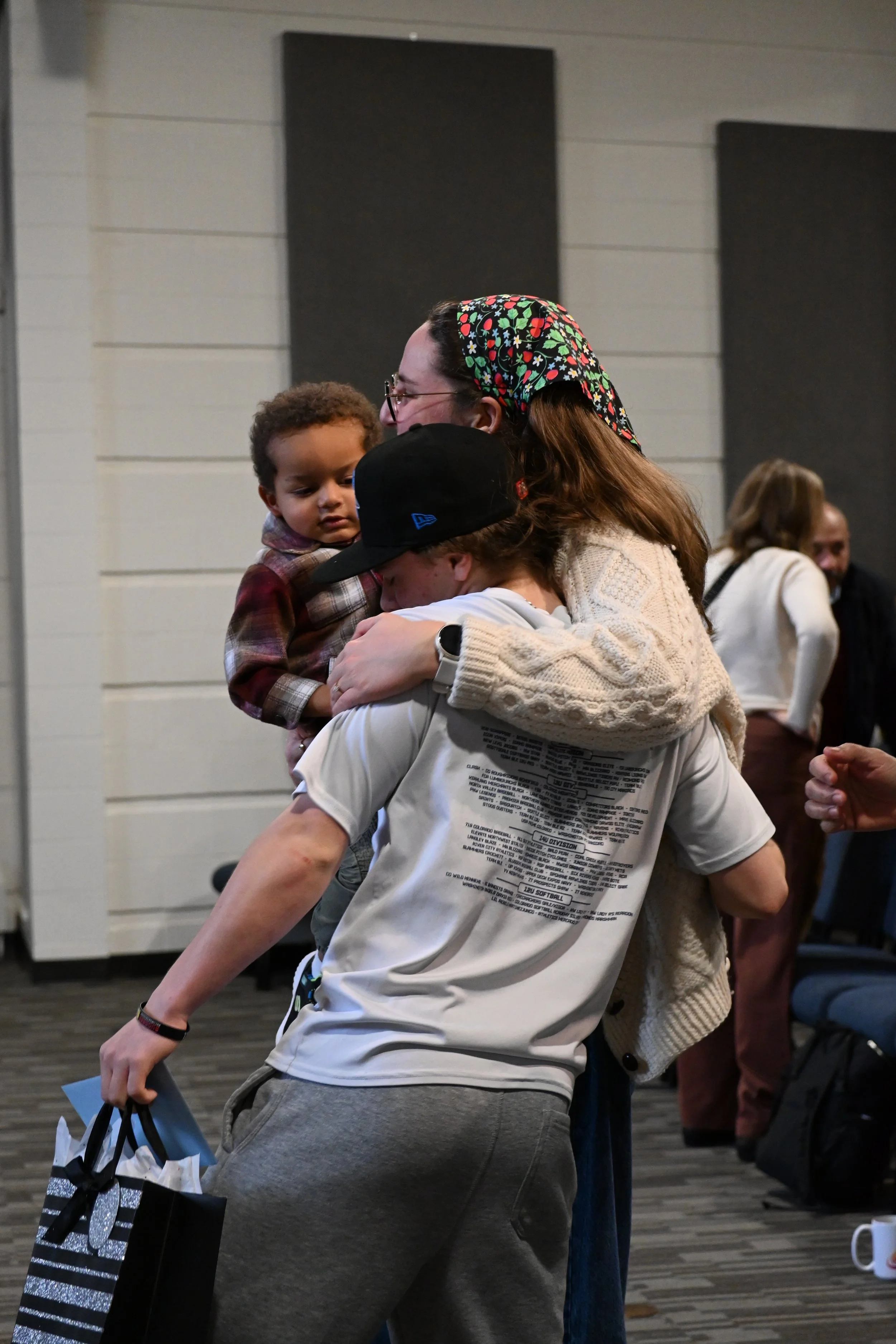 A woman hugging a man holding a young child, with people in the background in an indoor setting.
